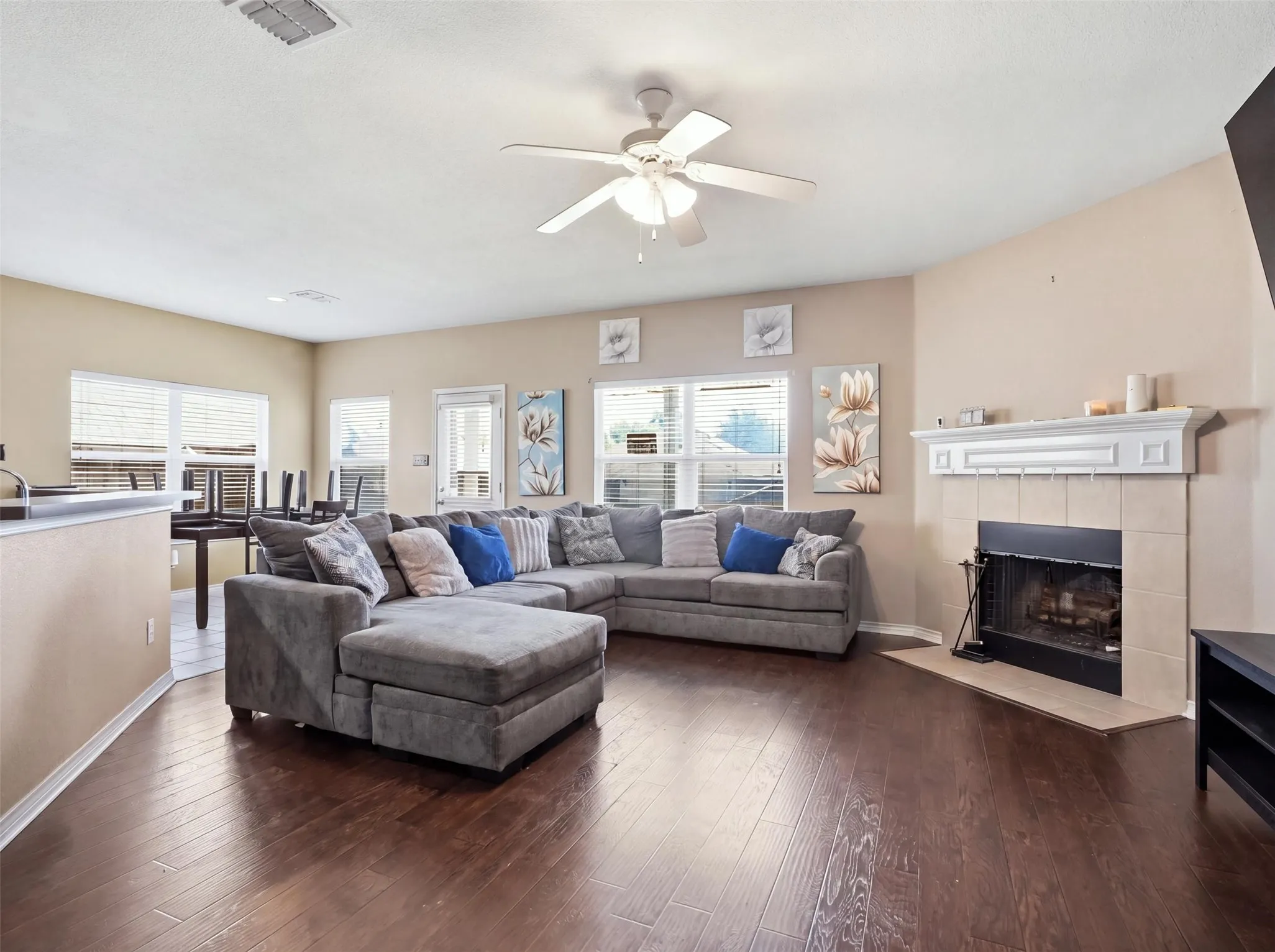 Spacious living area featuring wood-finish flooring, multiple windows with blinds, a ceiling fan, and a tiled fireplace with a mantel