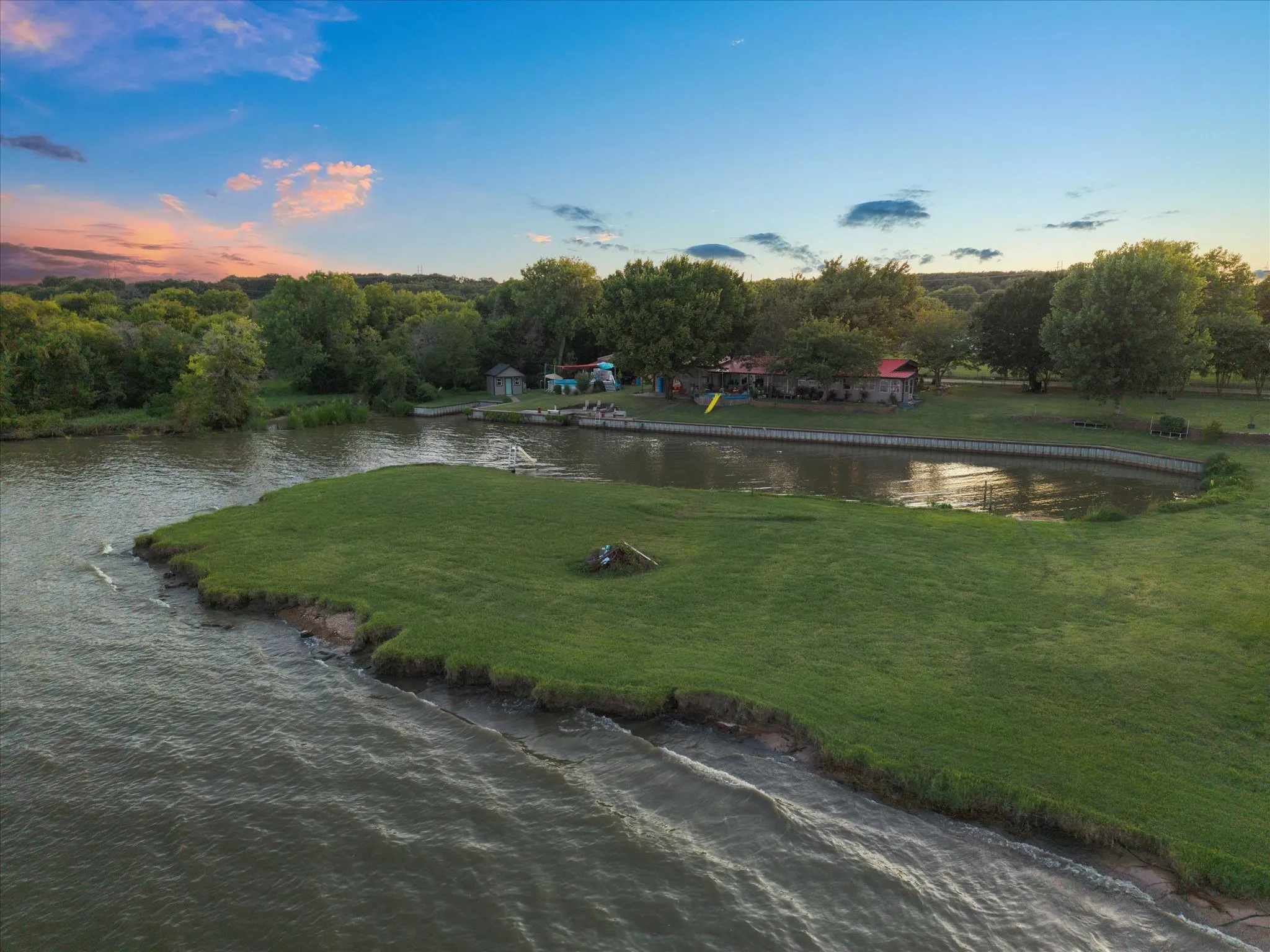 Waterfront property featuring a grassy peninsula, mature trees, and a retaining wall along the water's edge
