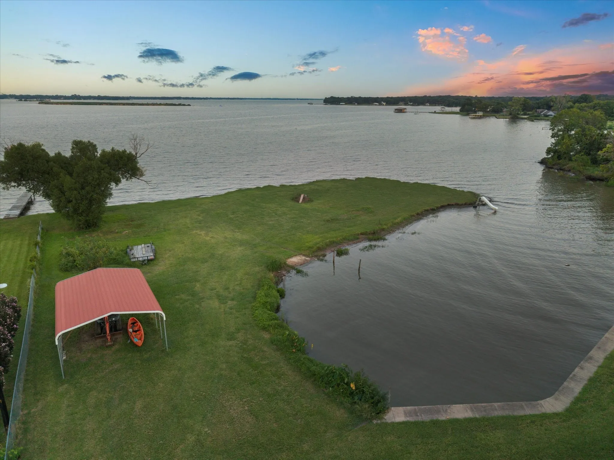 Expansive waterfront property featuring a manicured lawn, an extended pier, a red metal carport, and a concrete bulkhead