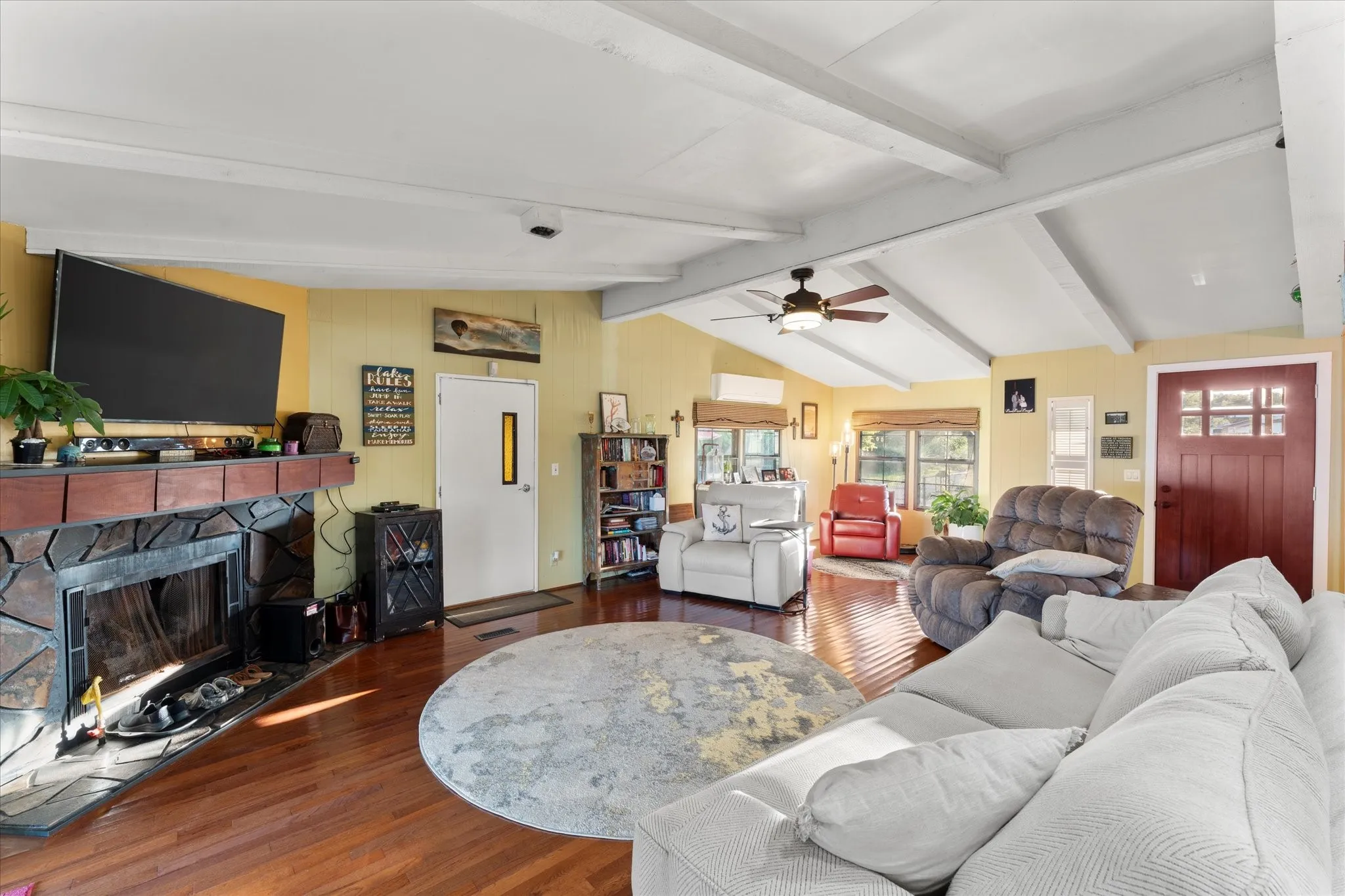 Vaulted ceiling with exposed beams, wood-finish flooring, stone-clad fireplace with mantel, ceiling fan, and red wood-paneled entry door
