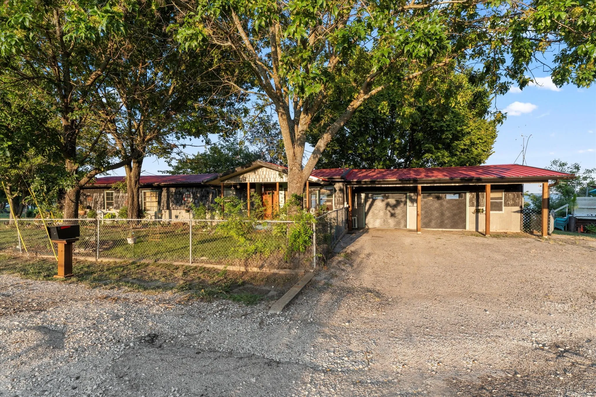 Red metal roof, gravel driveway, chain link fencing, multiple mature trees, and a covered carport