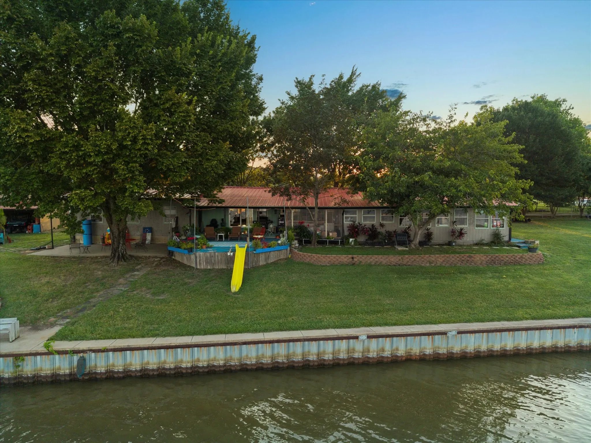 Waterfront property with a corrugated metal retaining wall, expansive green lawn, mature trees, and an elevated wooden deck