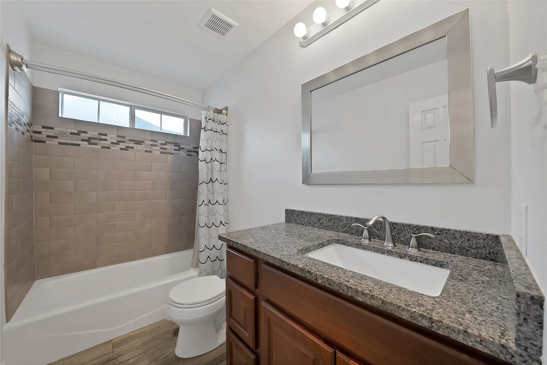 Wood-finish flooring, full-size bathtub, tiled shower surround with accent band, integrated vanity sink, and framed mirror