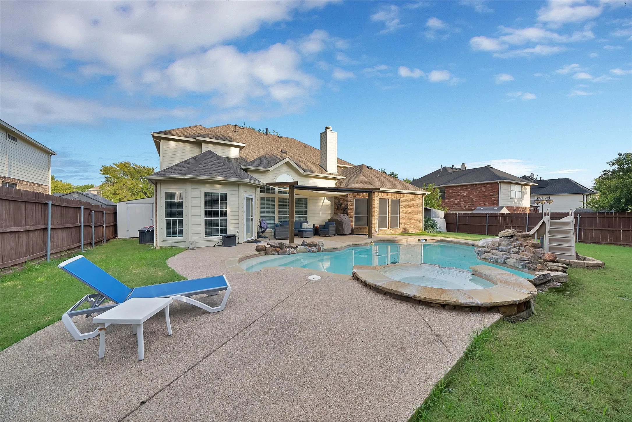 Expansive concrete patio surrounding a custom-shaped pool with an integrated spa, featuring an artificial rock waterfall and a pool slide