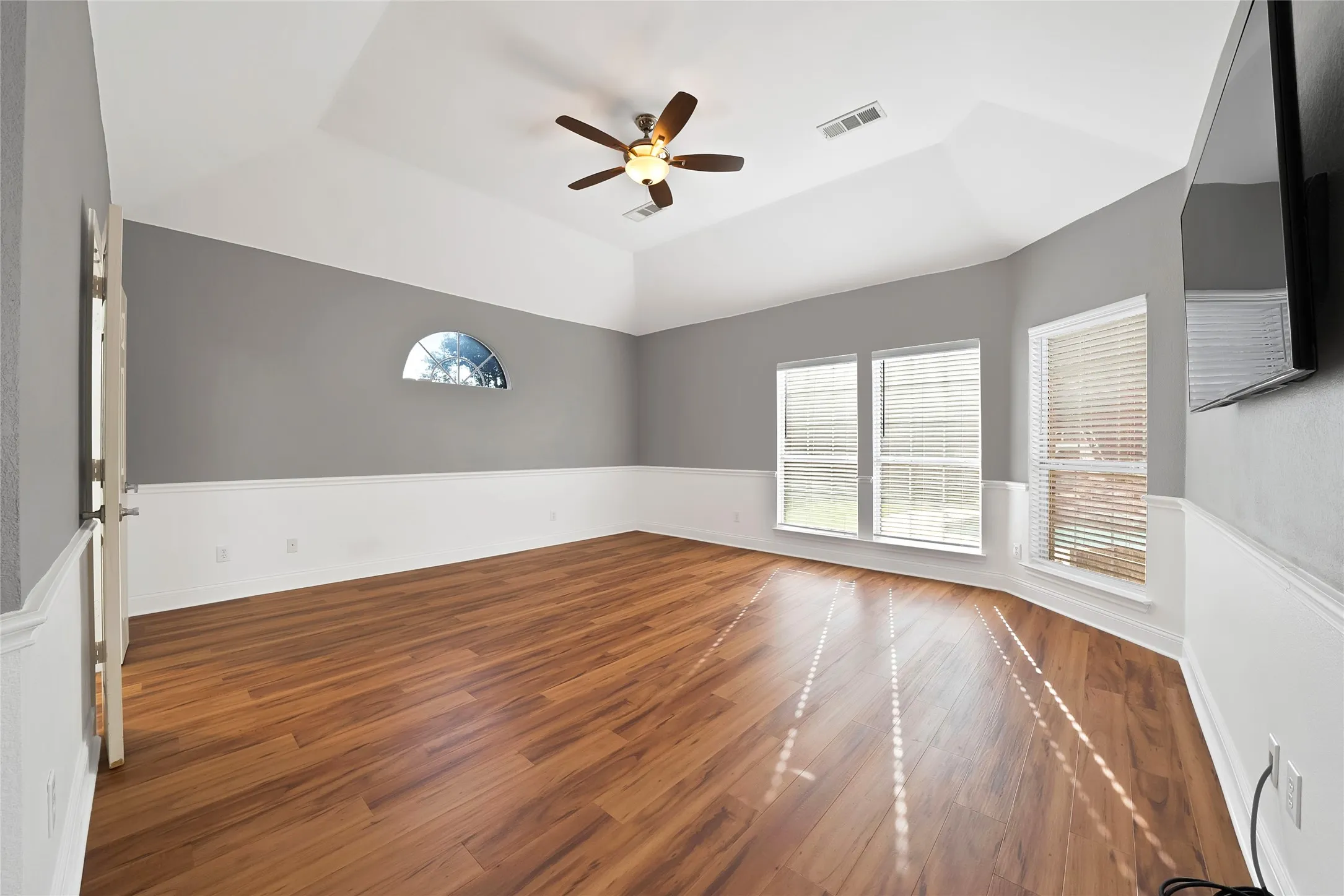 Vaulted ceiling with a ceiling fan, wood-finish flooring, half-round window, multiple windows with blinds, and two-tone wall paneling
