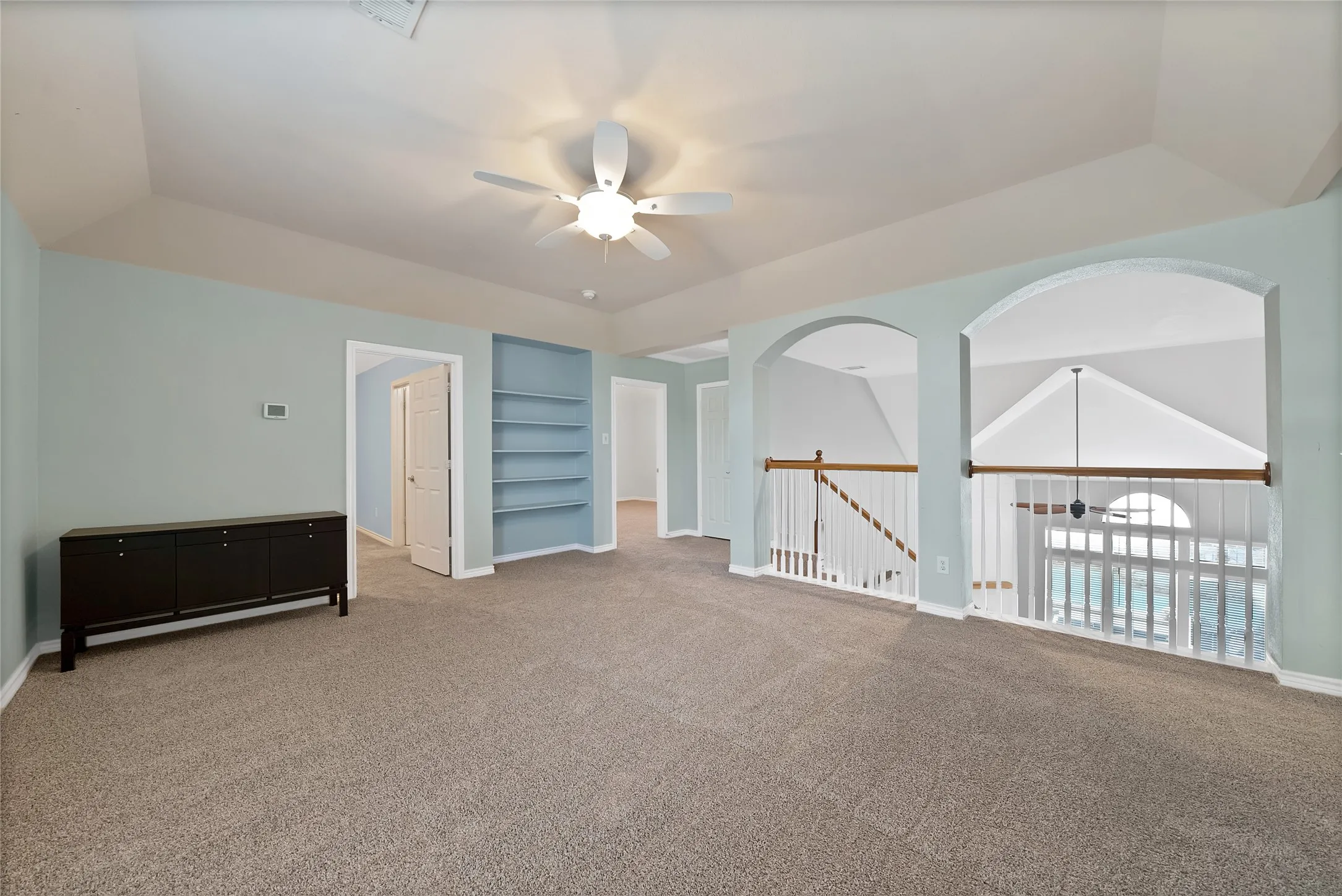 Tray ceiling with a ceiling fan, built-in shelving, two arched wall openings, white baluster railings, and neutral carpet flooring