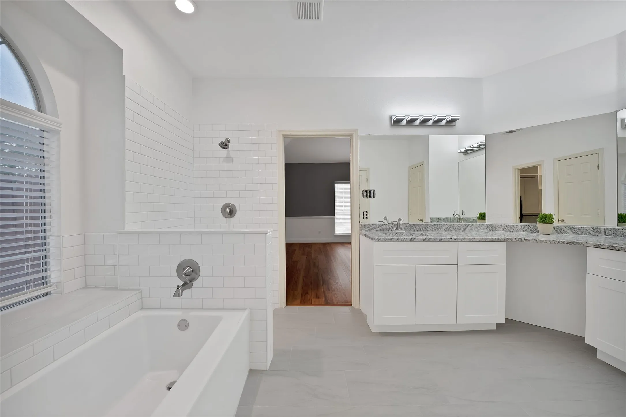 White subway tile shower surround, built-in bathtub, dual sink vanity with granite-style countertop, and tile flooring