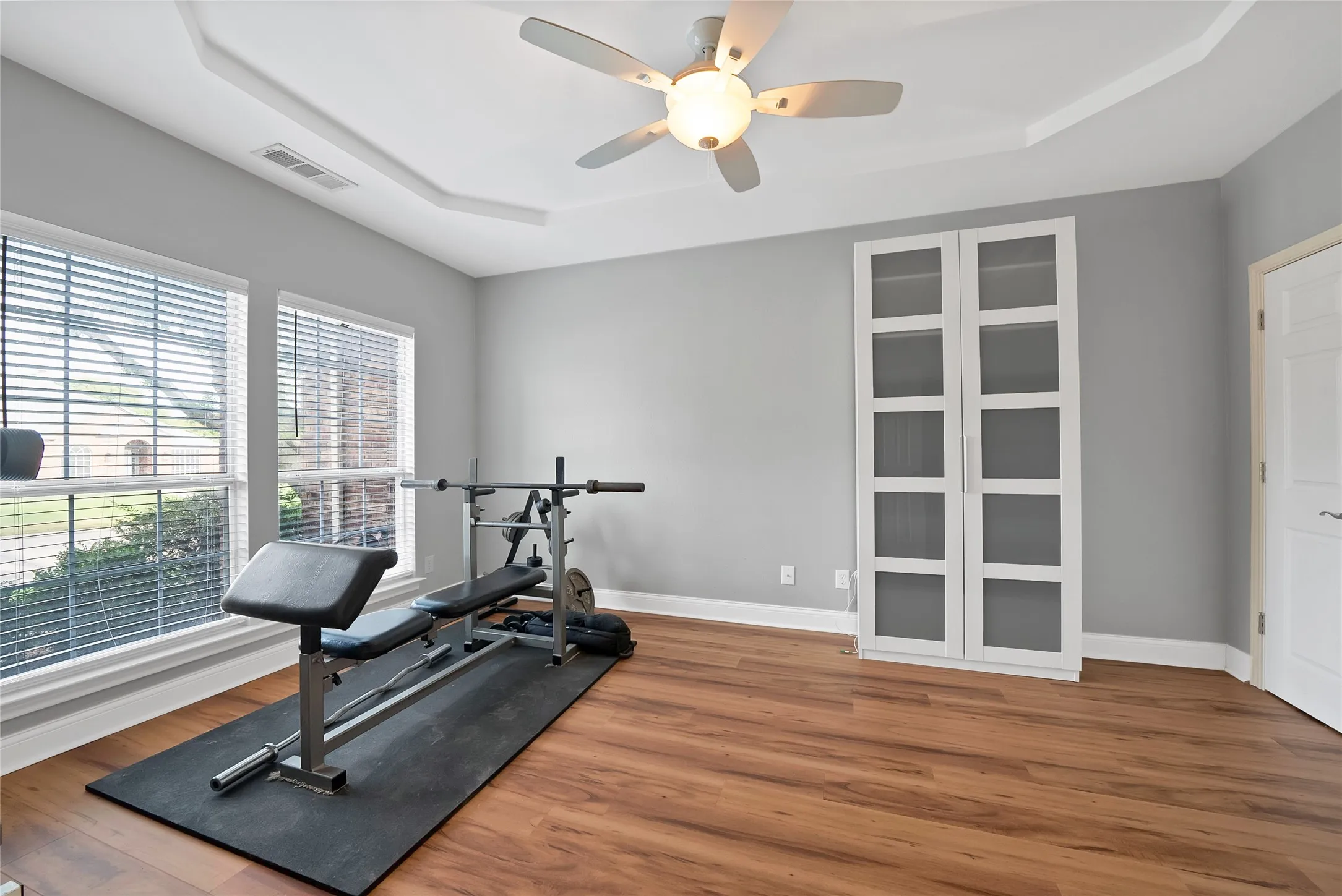 Tray ceiling with a ceiling fan, wood-finish flooring, and white built-in cabinetry with glass panel inserts