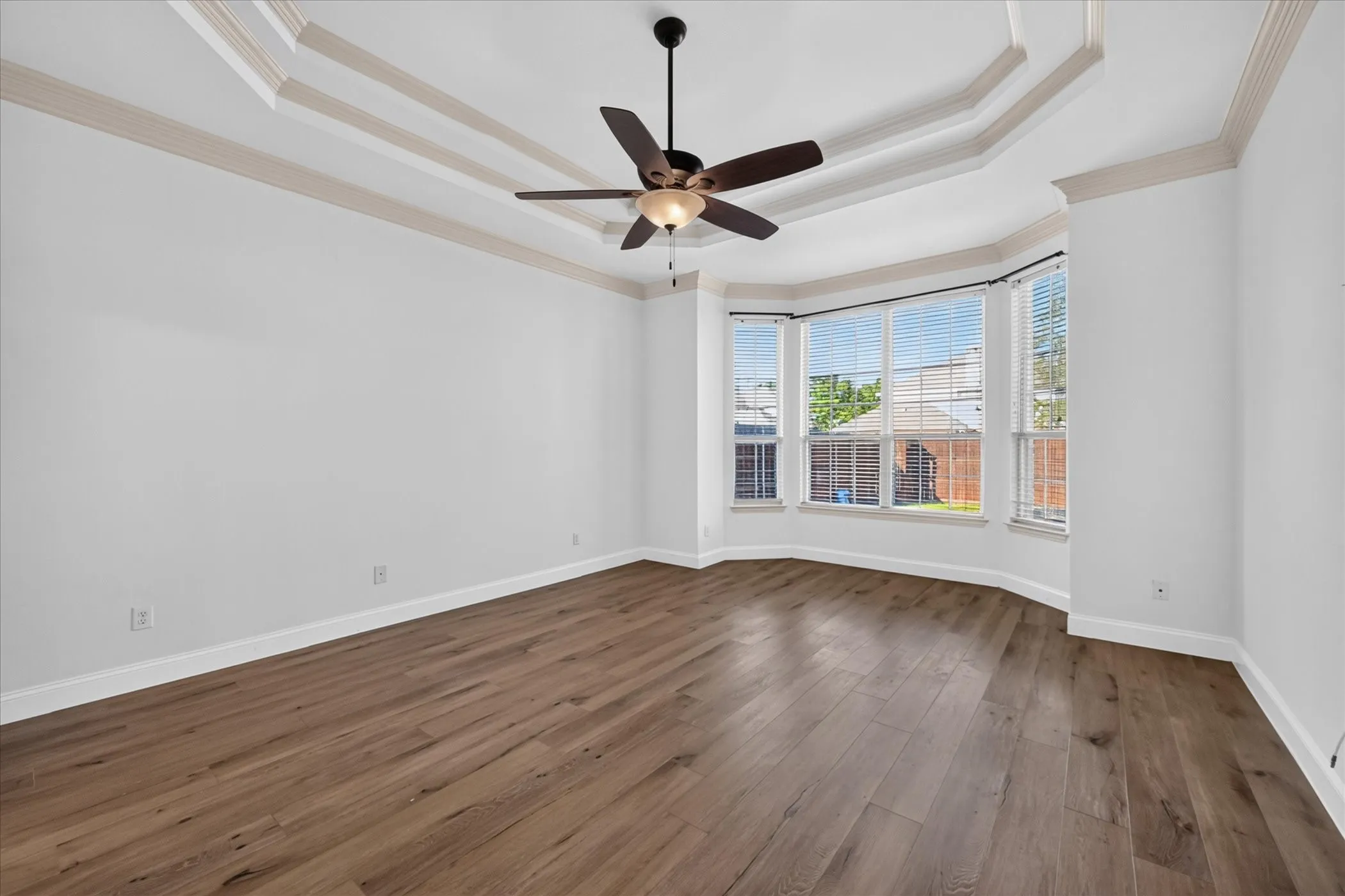 Tray ceiling with decorative molding, wood-finish flooring, bay windows, and a ceiling fan with integrated lighting