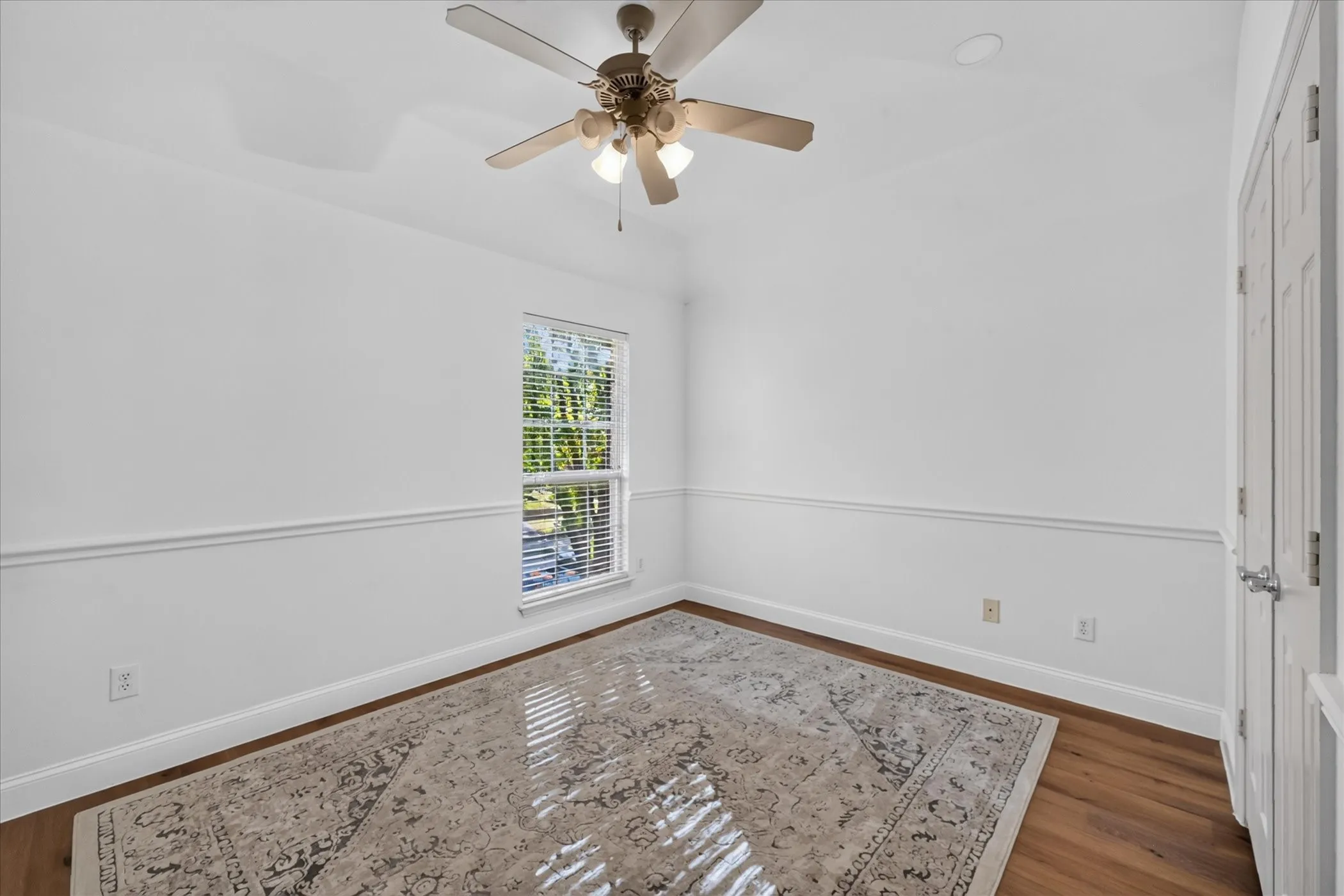 Recessed ceiling with a ceiling fan, hardwood flooring, natural light through a window with blinds, and white interior walls featuring chair rail molding