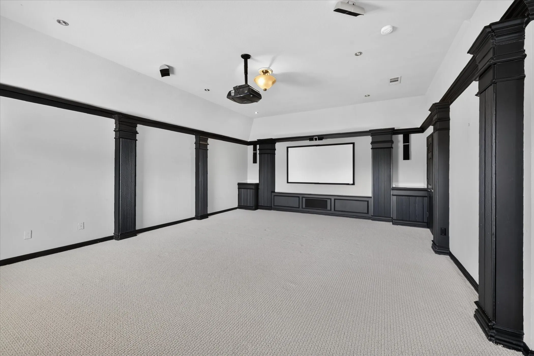Dedicated media room featuring recessed ceiling lighting, a ceiling-mounted projector, built-in cabinetry with display shelving, large projection screen, black architectural framing elements, and neutral carpet flooring throughout