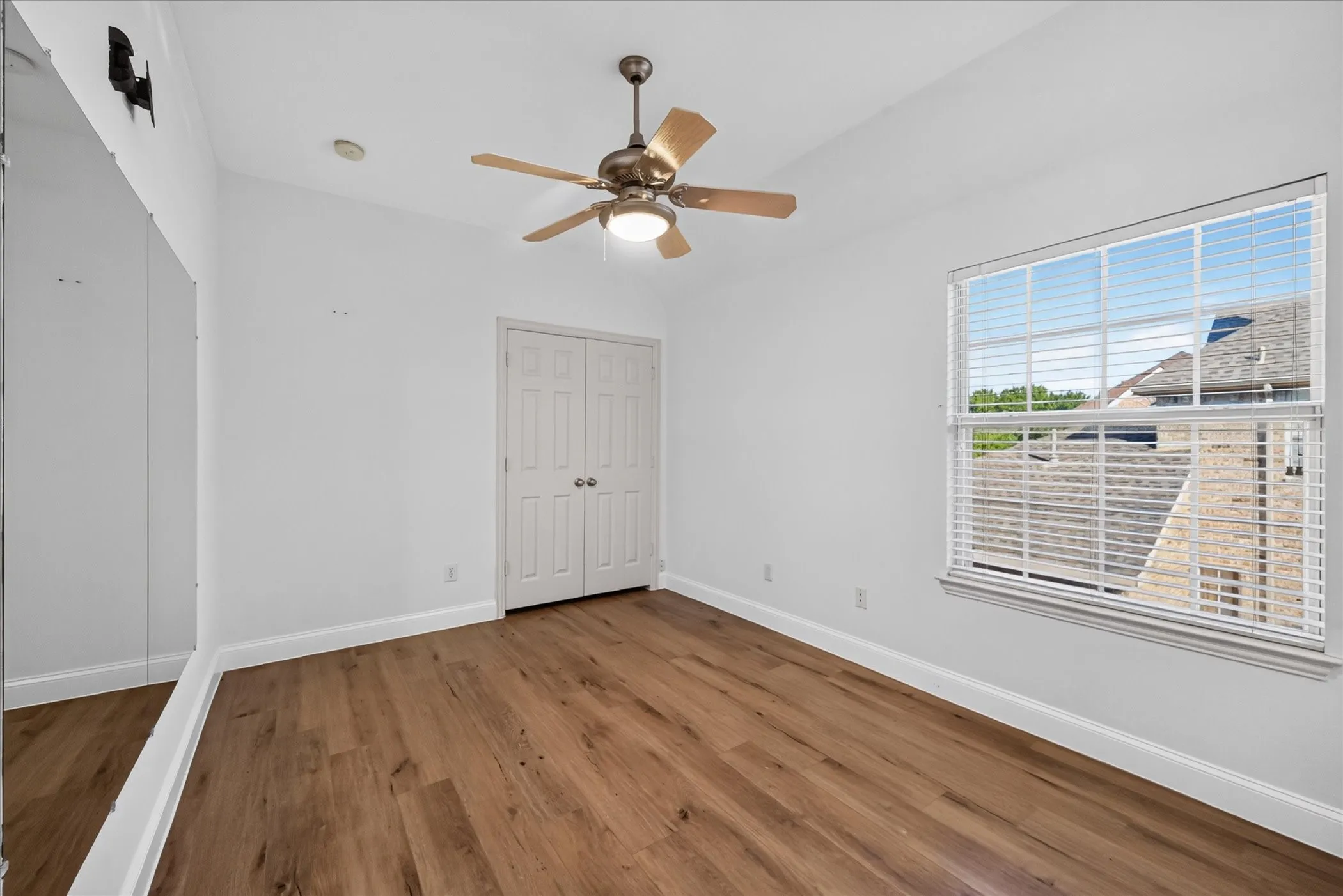Wood-finish flooring, ceiling fan with integrated lighting, window with horizontal blinds, built-in bi-fold closet doors, and a full-height wall mirror
