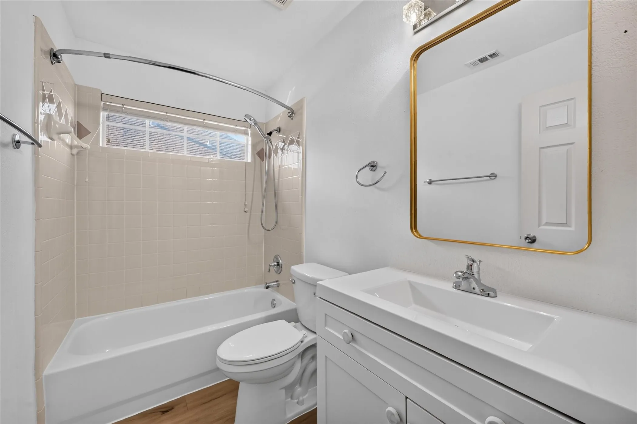 White vanity with integrated sink, rectangular gold-framed mirror, wood-finish flooring, and a shower-tub combination with light-toned wall tile