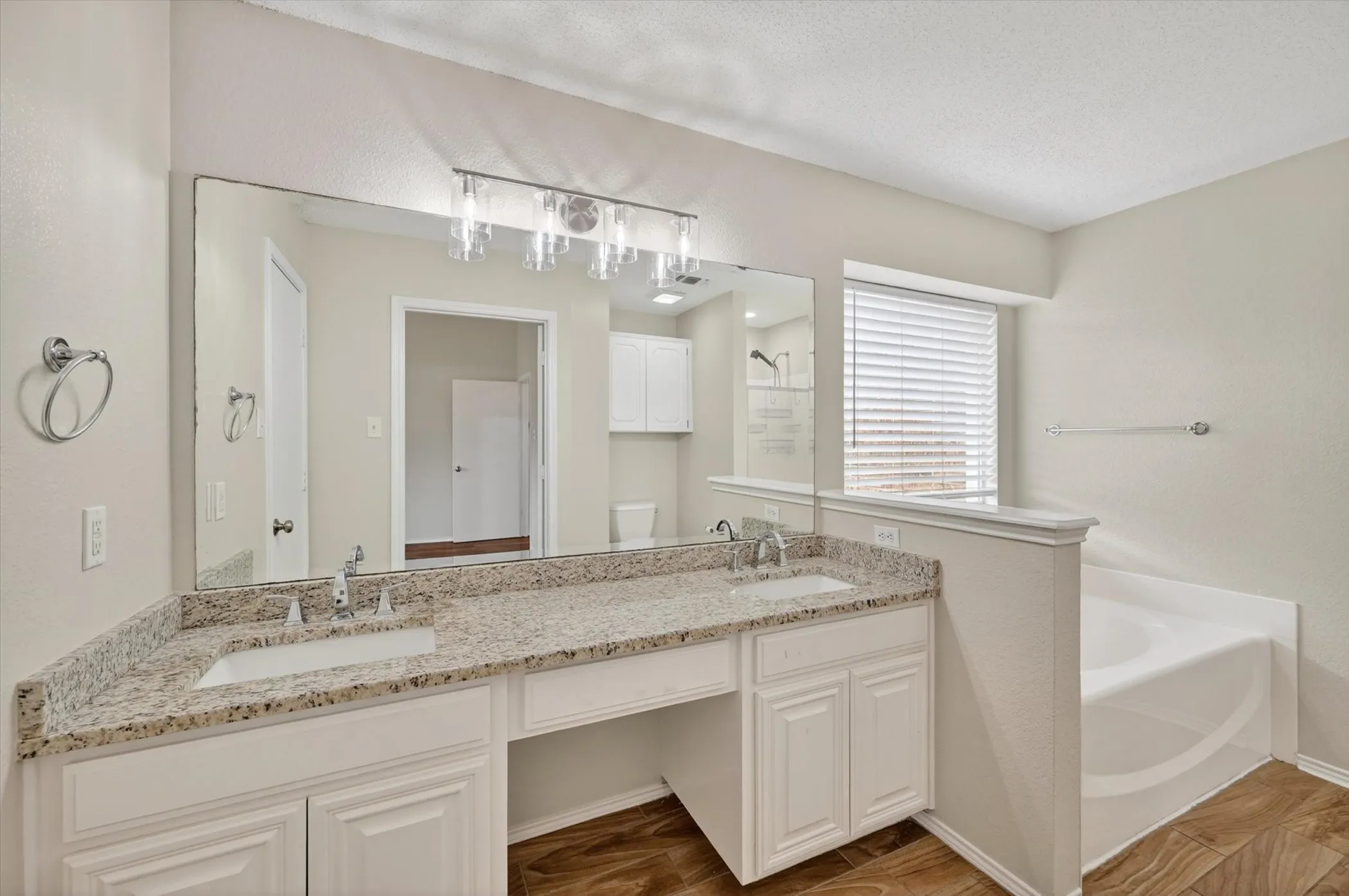 Dual vanity with integrated sinks, speckled countertop, decorative light fixture, built-in bathtub, and wood-finish flooring