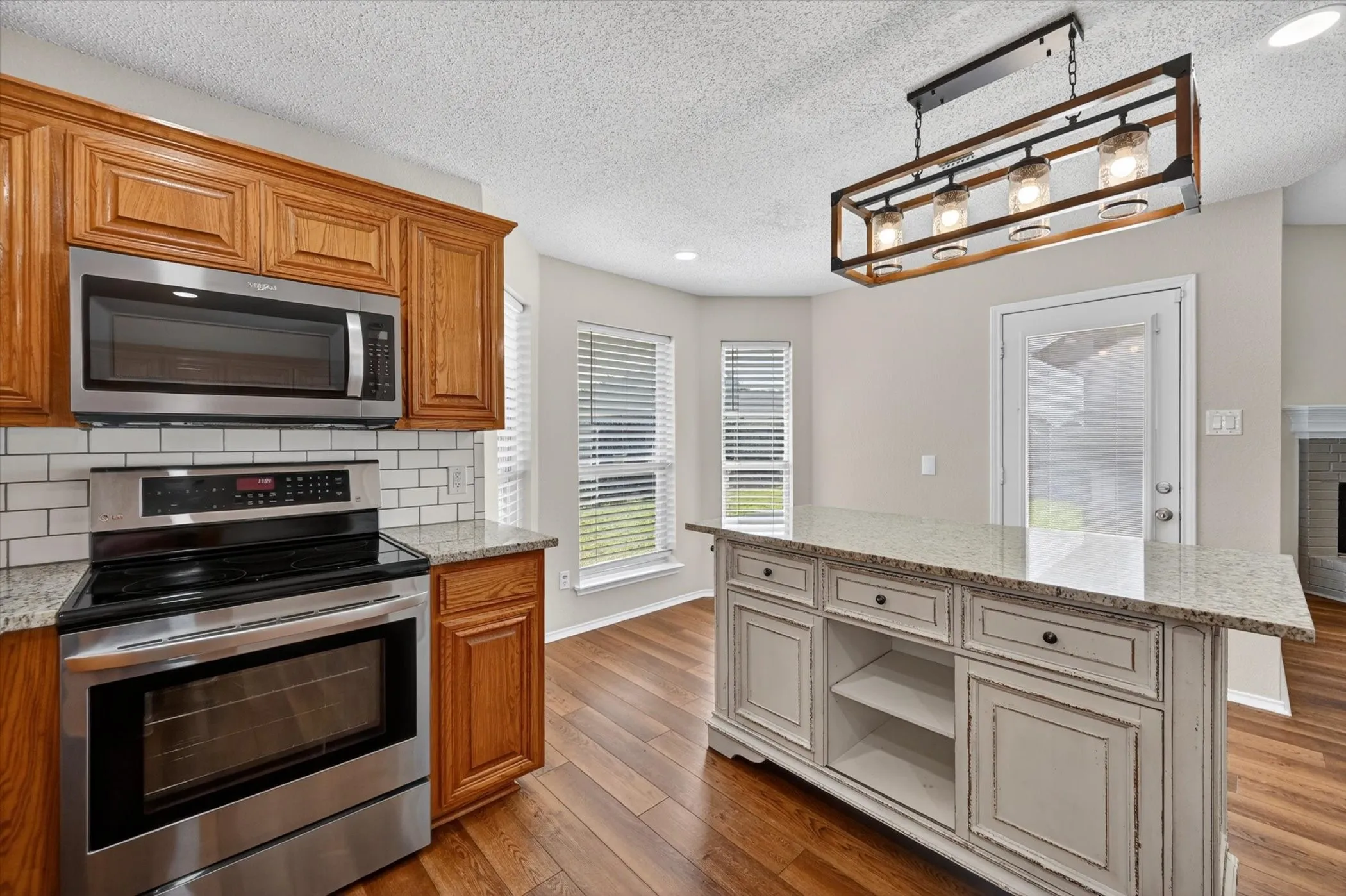 Wood-finish flooring, island with granite-style countertop, stainless steel appliances, white subway tile backsplash, and wood cabinetry