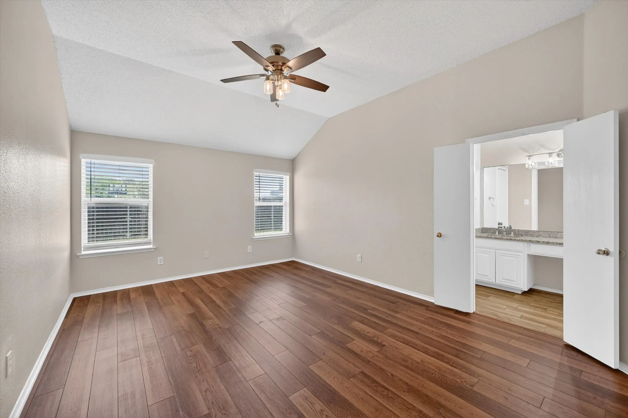 Vaulted ceiling, wood-finish flooring, two windows with blinds, ceiling fan with light fixture, and a vanity with a mirror and built-in cabinetry