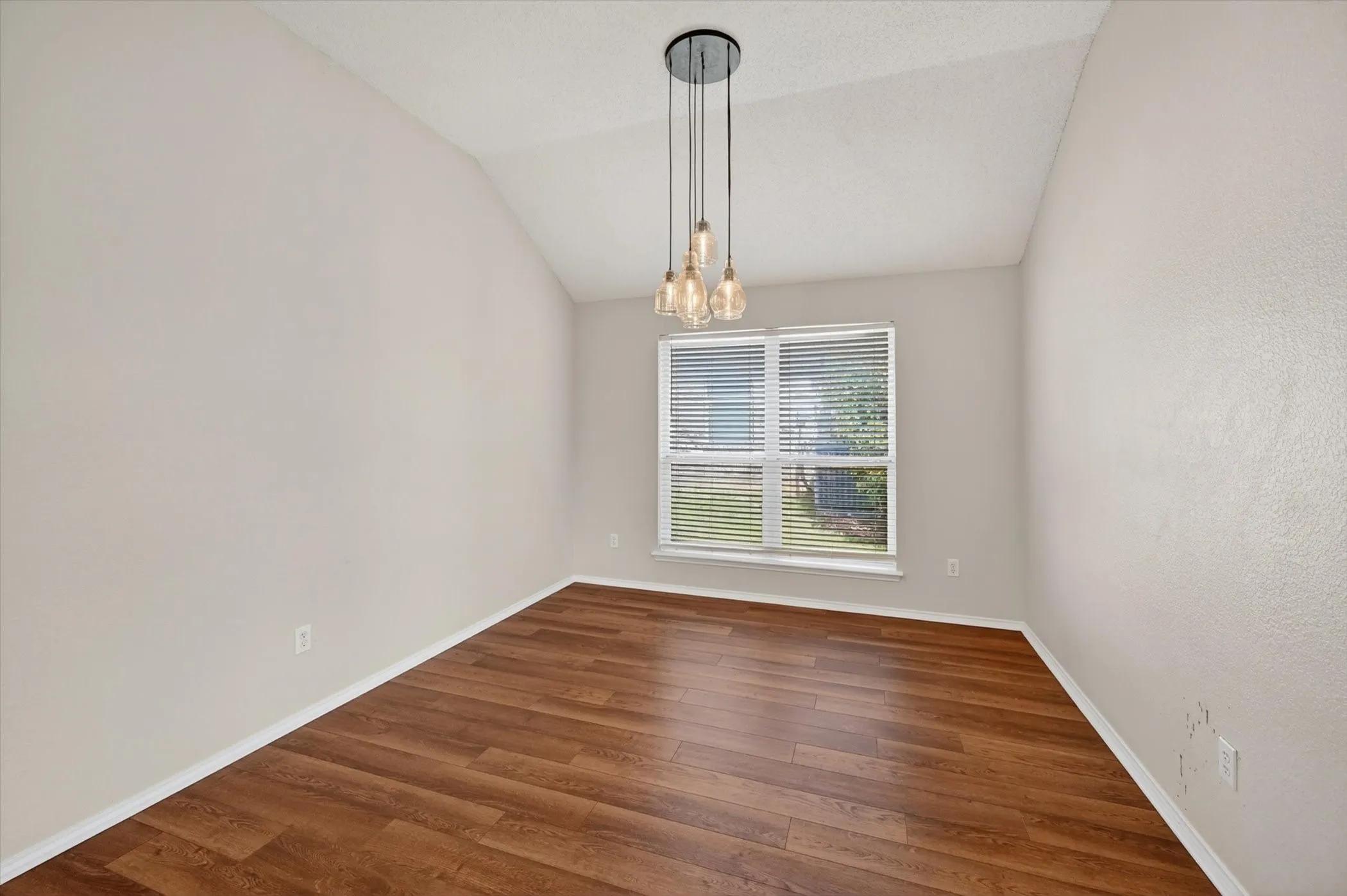 Vaulted ceiling, wood-finish flooring, multiple pane window, built-in window blinds, and a multi-bulb hanging fixture