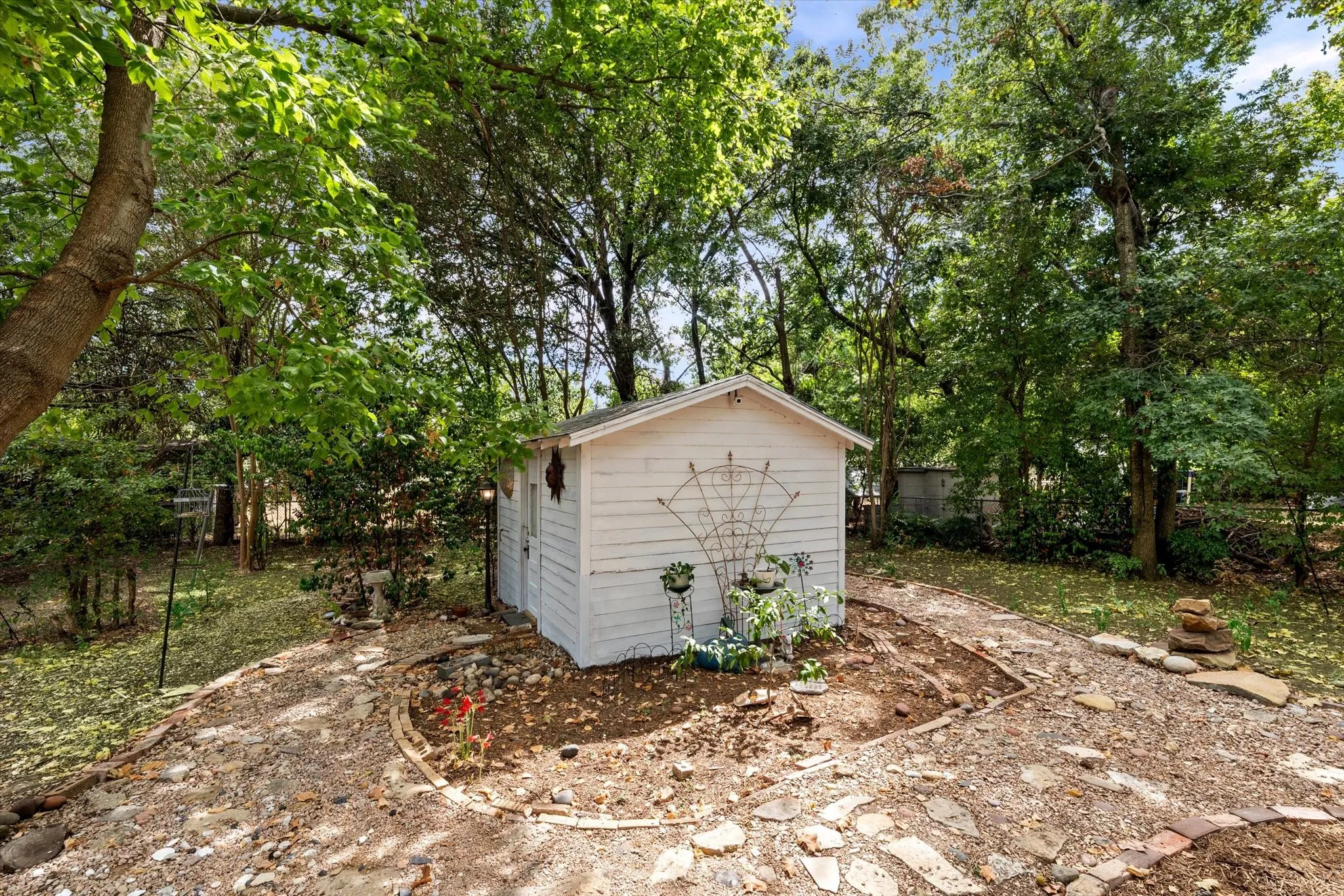 Backyard storage shed with white horizontal siding and a gable roof, surrounded by mature trees and a stone pathway