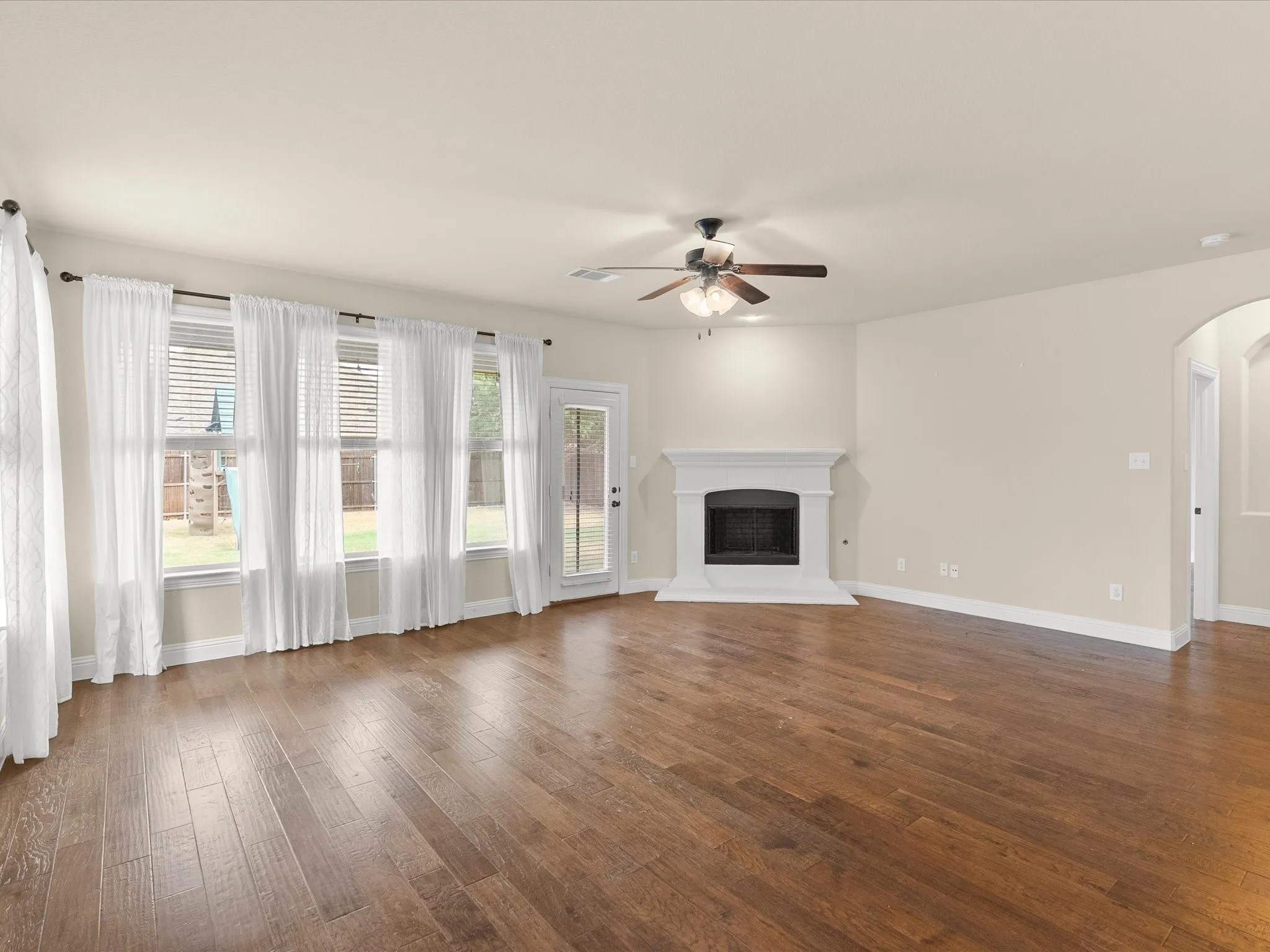 Spacious room featuring wood-finish flooring, multiple windows, a ceiling fan, and a fireplace with a white mantle