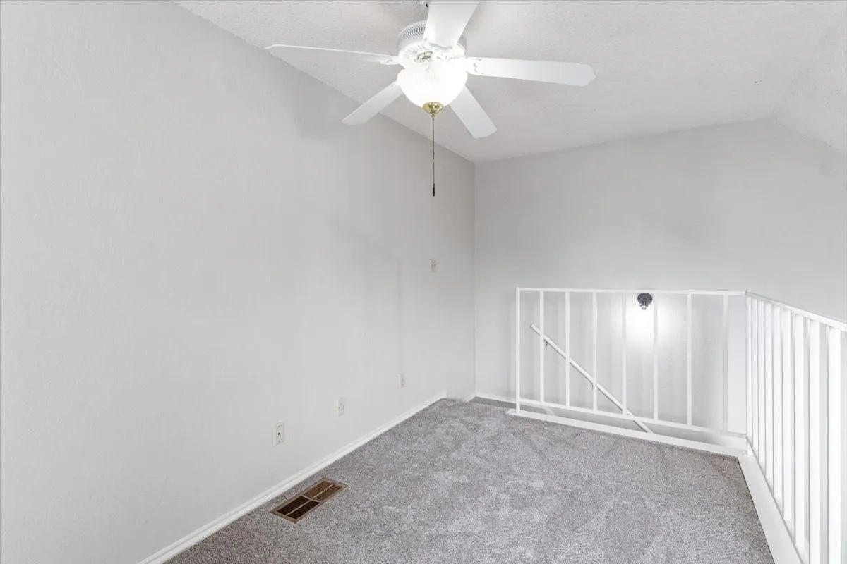 Carpeted room featuring a ceiling fan with light, white painted walls, and a white railing