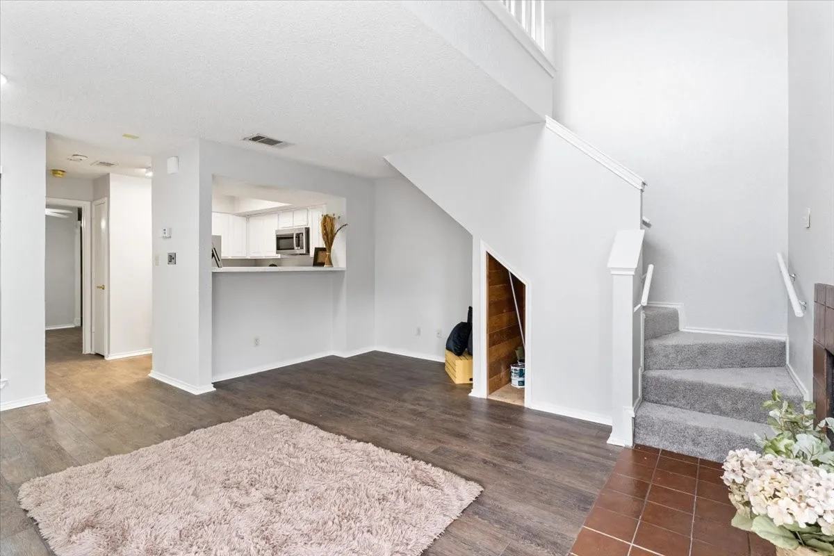 Open-concept living area featuring wood-finish flooring, a pass-through kitchen with white cabinetry, a carpeted staircase with a white railing, and an under-stair storage area with wood paneling