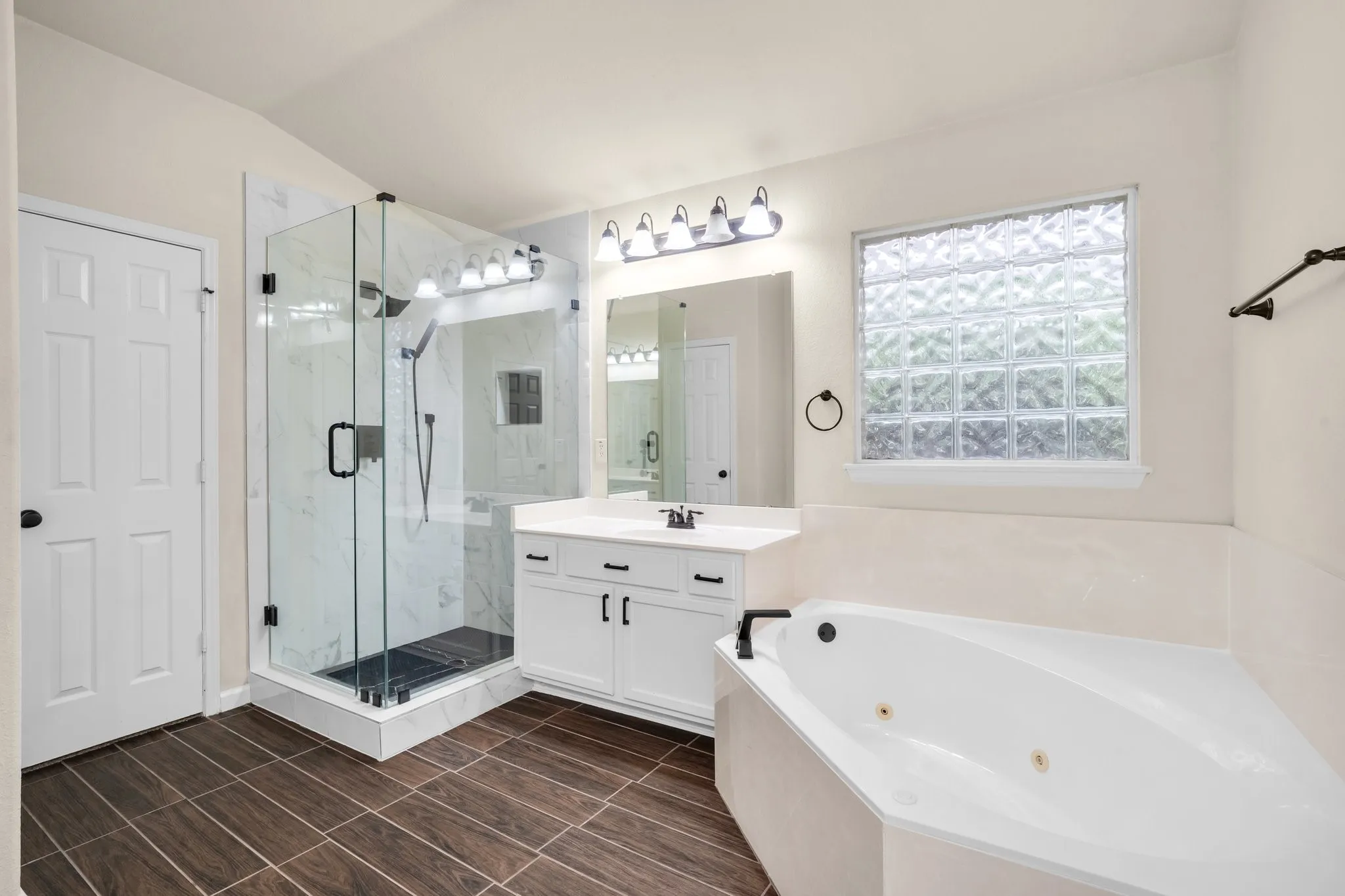 Wood-finish flooring, glass-enclosed shower with tiled walls, white vanity with integrated sink, contemporary light fixture, and jetted corner tub