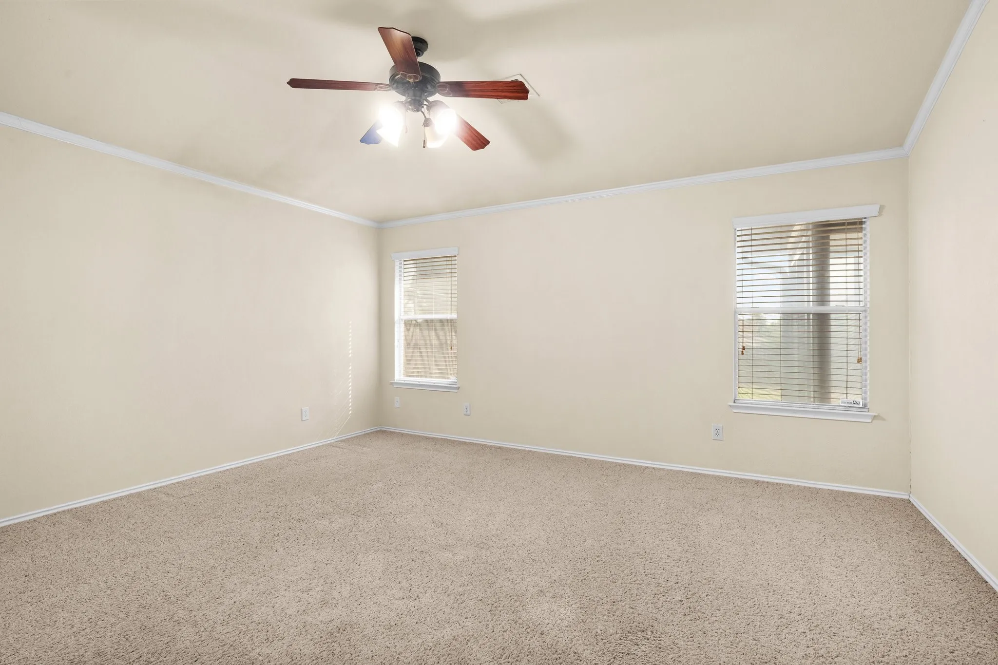 Spacious room featuring neutral carpeting, crown molding, two windows with blinds, and a ceiling fan with integrated lighting