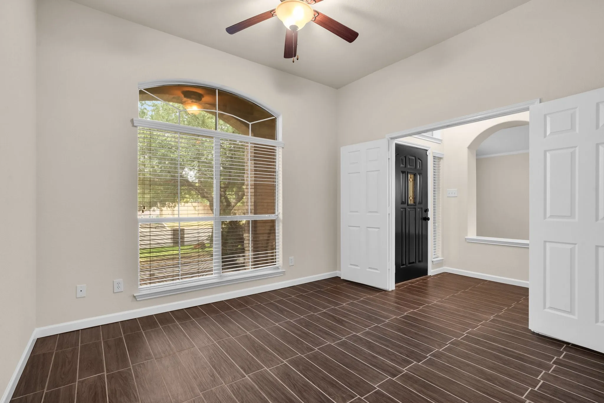 Arched window with blinds, wood-finish flooring, ceiling fan, white paneled double doors, and a black front door with decorative glass