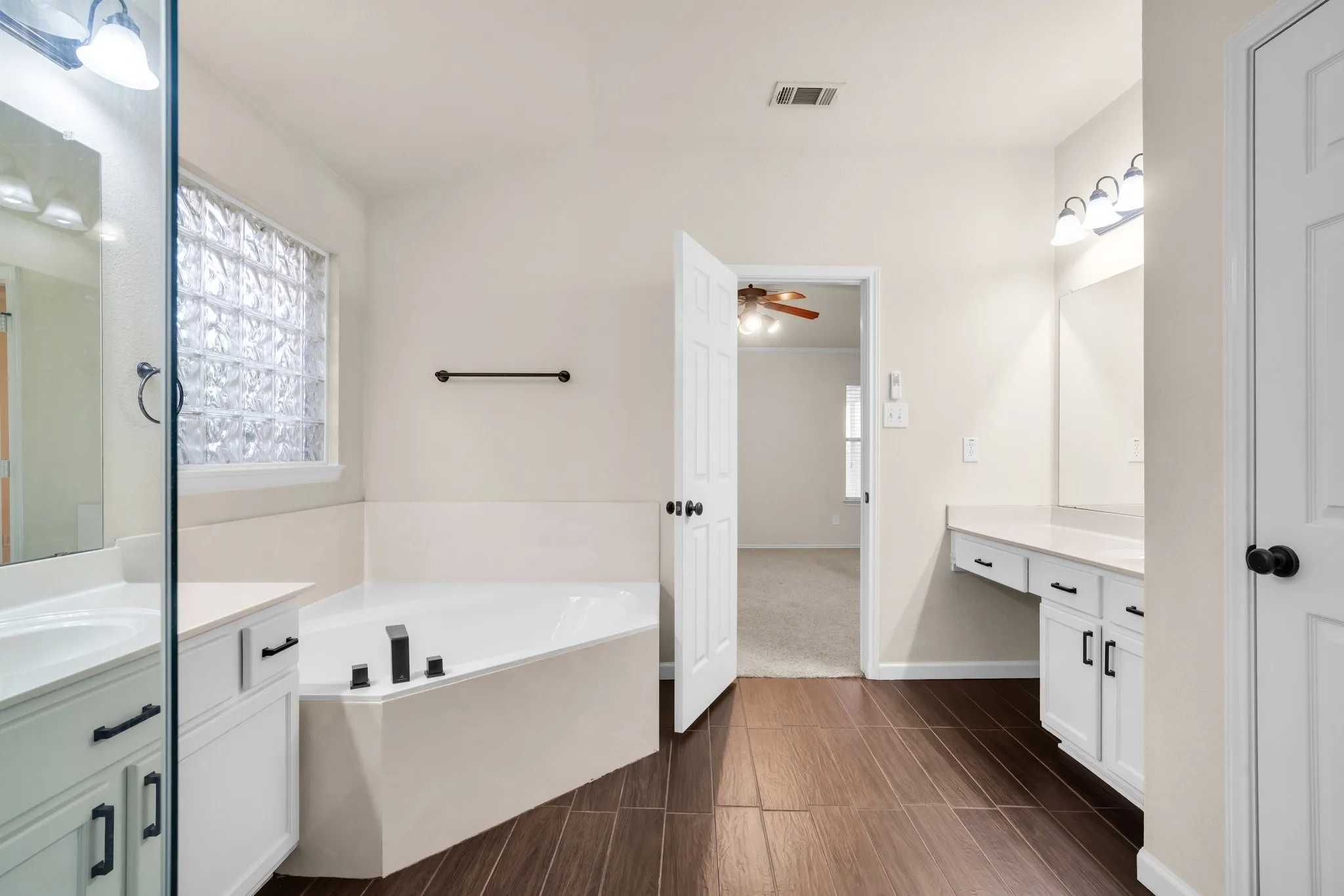 Wood-finish flooring, corner soaking tub, dual vanities with white cabinetry, and a glass block window