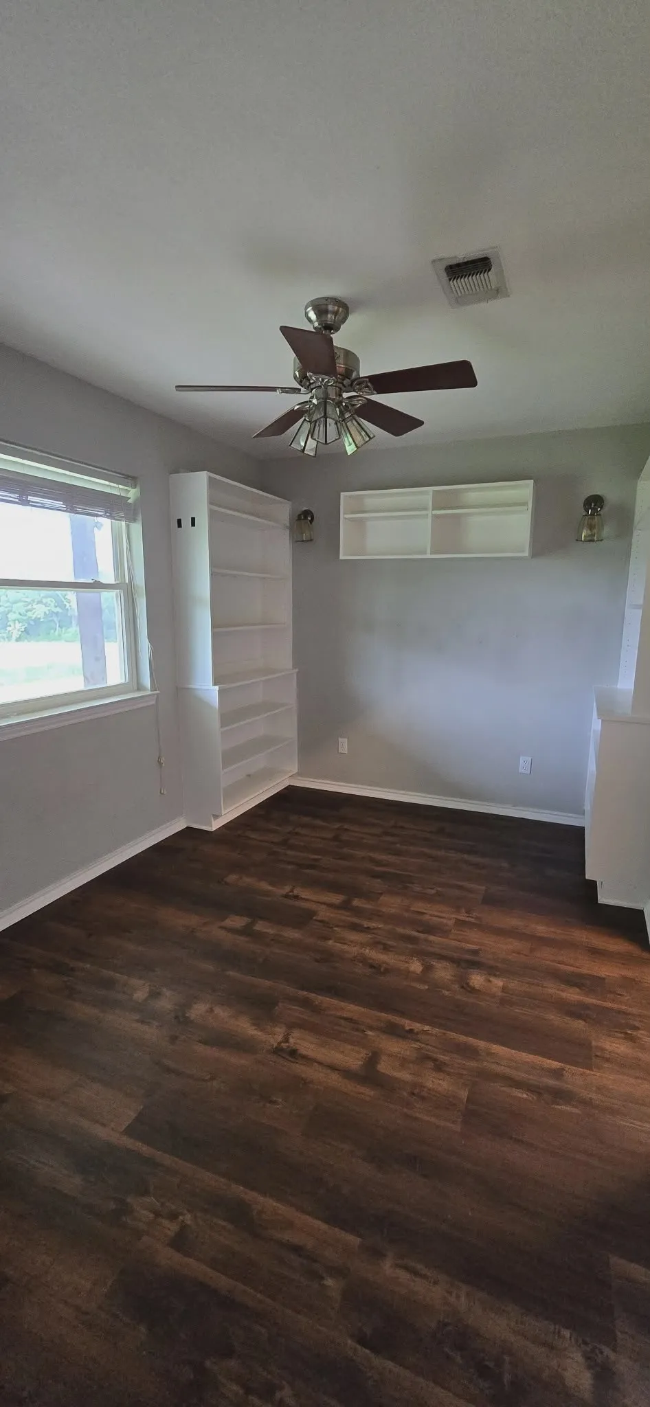 Wood-finish flooring, built-in shelving, windows providing natural light, ceiling fan with integrated lighting, and wall-mounted sconces