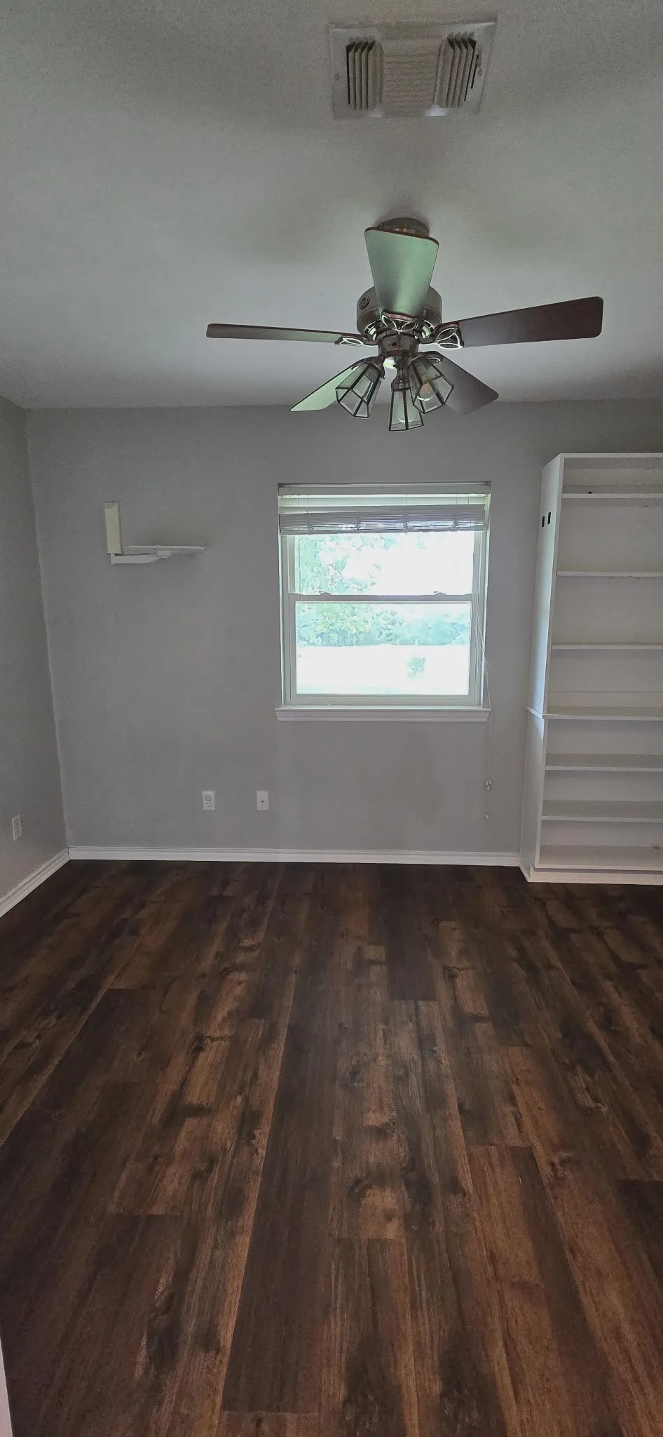Wood-finish flooring, ceiling fan with integrated lighting, built-in shelving, white window blinds, and light gray wall paint