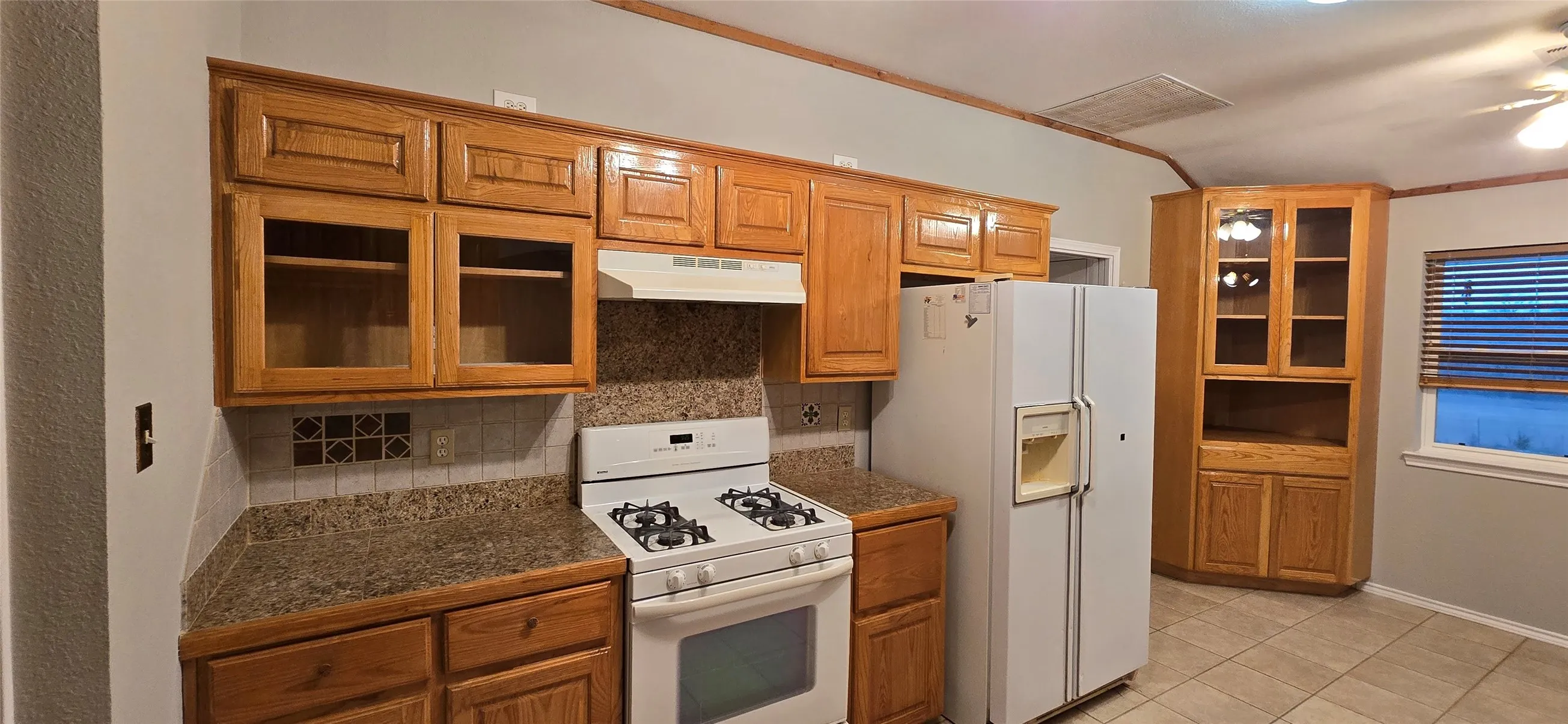Oak wood cabinetry with glass-front accents, solid surface countertops, tile backsplash with decorative inserts, and ceramic tile flooring