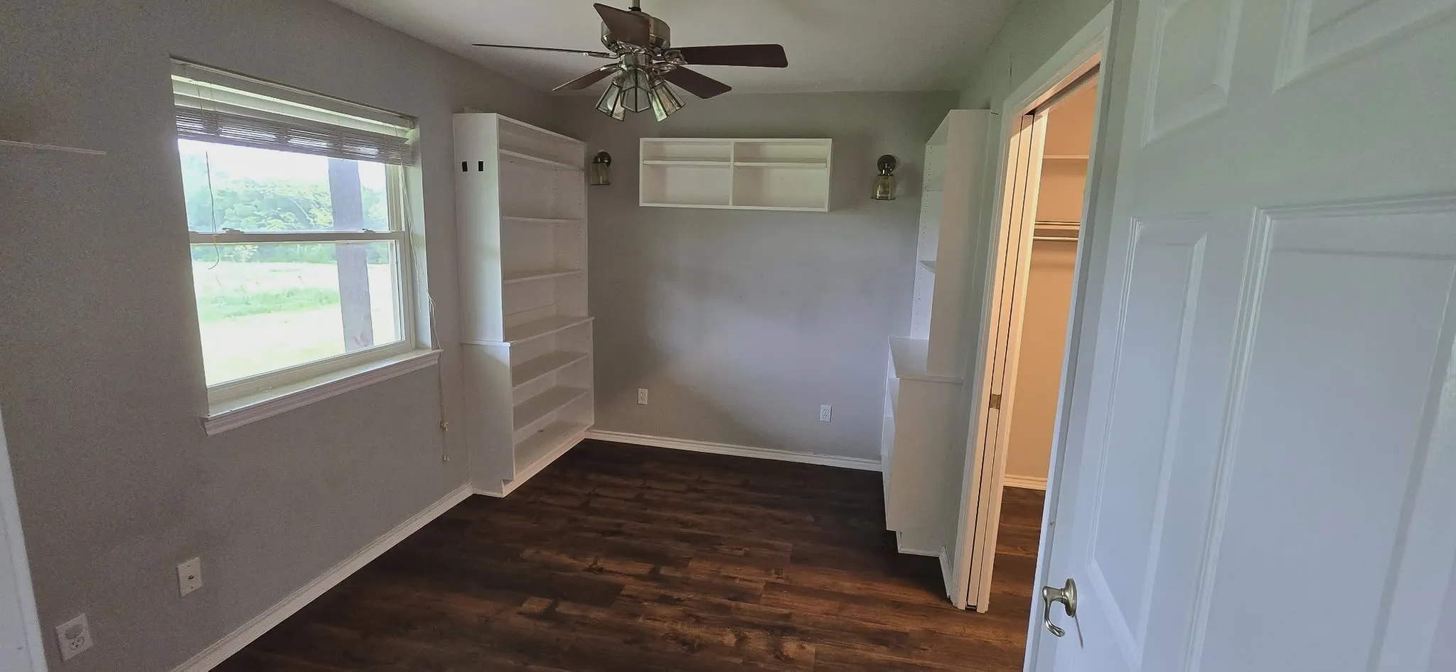 Wood-finish flooring, built-in shelving, ceiling fan with integrated lighting, and a window with blinds