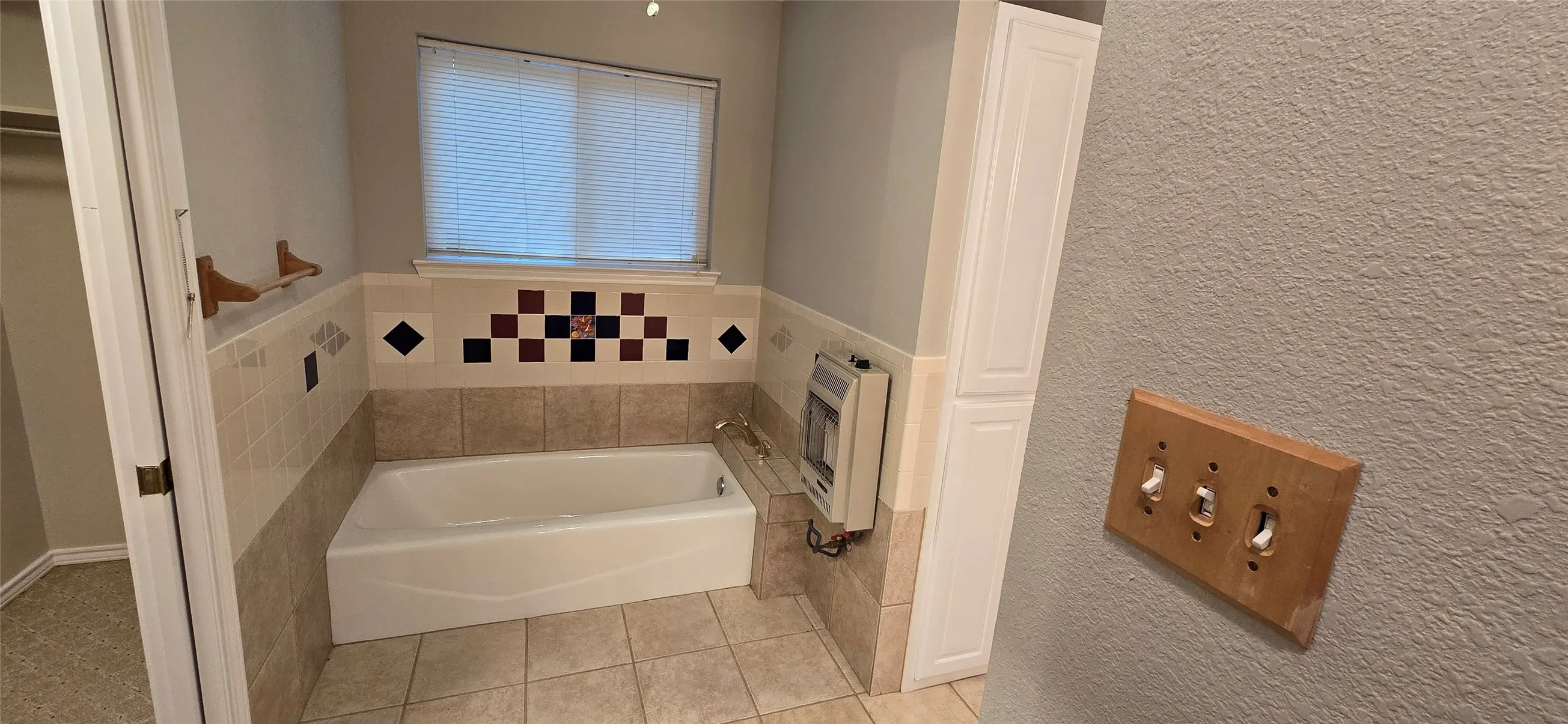 White bathtub with tile surround, window with blinds, and built-in white cabinetry
