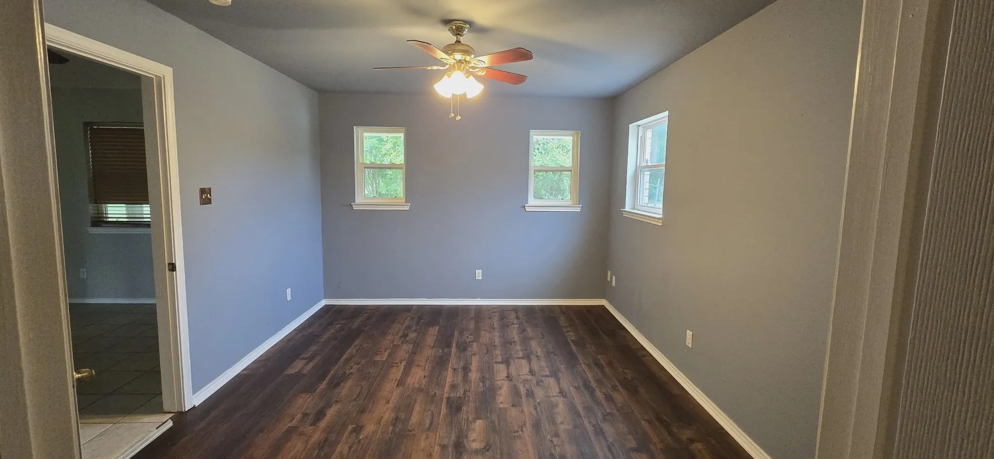 Wood-finish flooring with light gray walls, featuring multiple windows and a ceiling fan