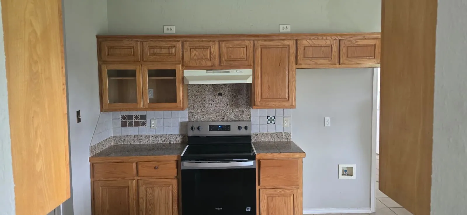 Wood cabinetry with glass-front accents, granite-look countertops, tile backsplash, and a modern stainless steel range