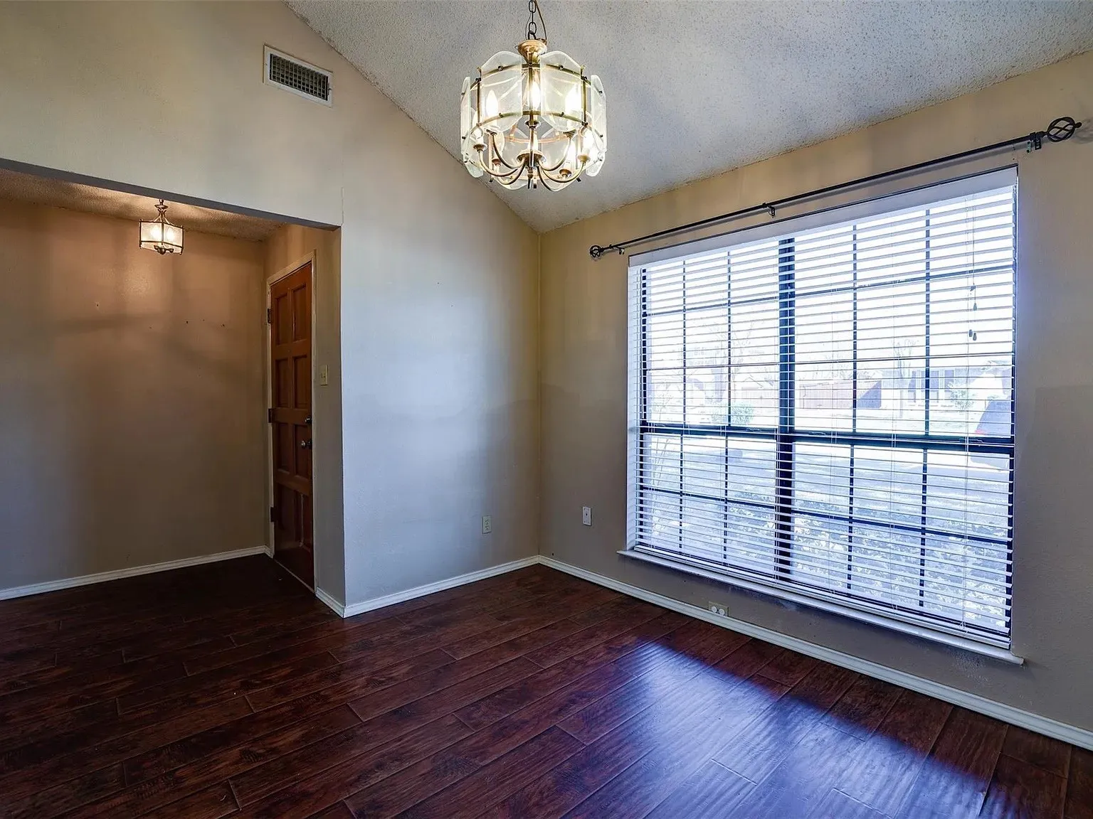 Vaulted ceiling with an ornate chandelier, expansive window with horizontal blinds, rich wood-finish flooring, and a paneled entry door