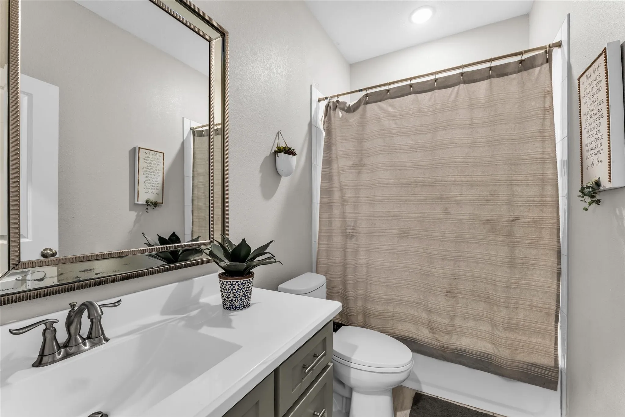 Spa-inspired bathroom featuring a framed mirror with integrated lighting, white ceramic sink with polished chrome faucet and handles, white subway tile surround, neutral linen shower curtain with metal rod, white porcelain toilet, and wood-finish vanity cabinetry with ample storage