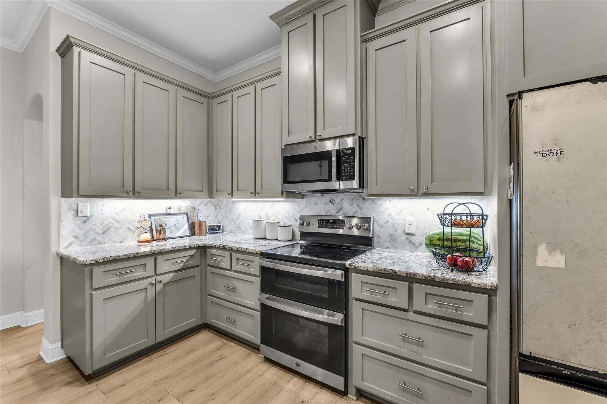 Gray cabinetry with crown molding, light-toned wood-finish flooring, stainless steel electric range, stainless steel microwave, and a tiled backsplash