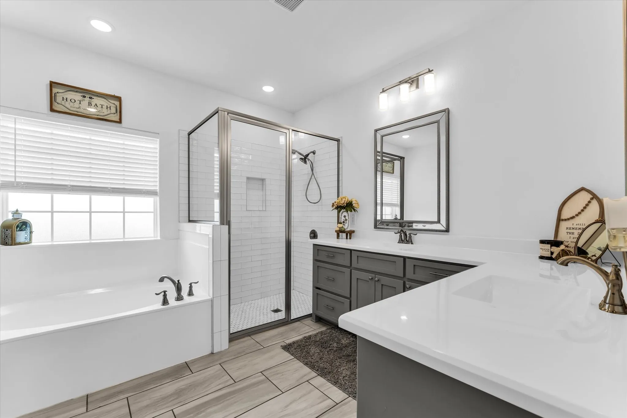 Spacious bathroom featuring a built-in soaking tub, glass-enclosed shower with subway tile, wood-finish tile flooring, and a double vanity with grey cabinetry and white countertops