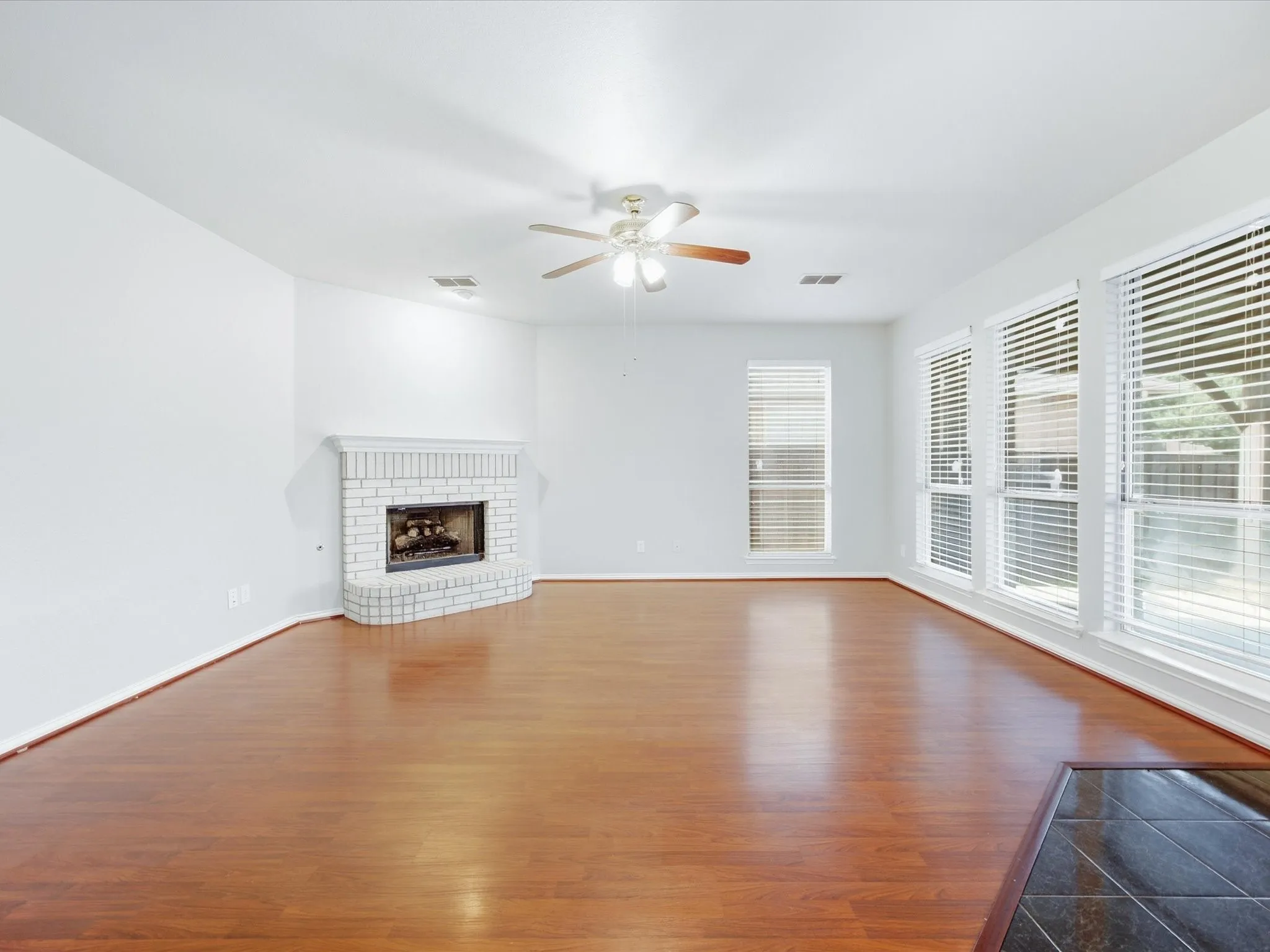 Expansive interior with wood-finish flooring, a brick-surround fireplace, multiple large windows, and a ceiling fan