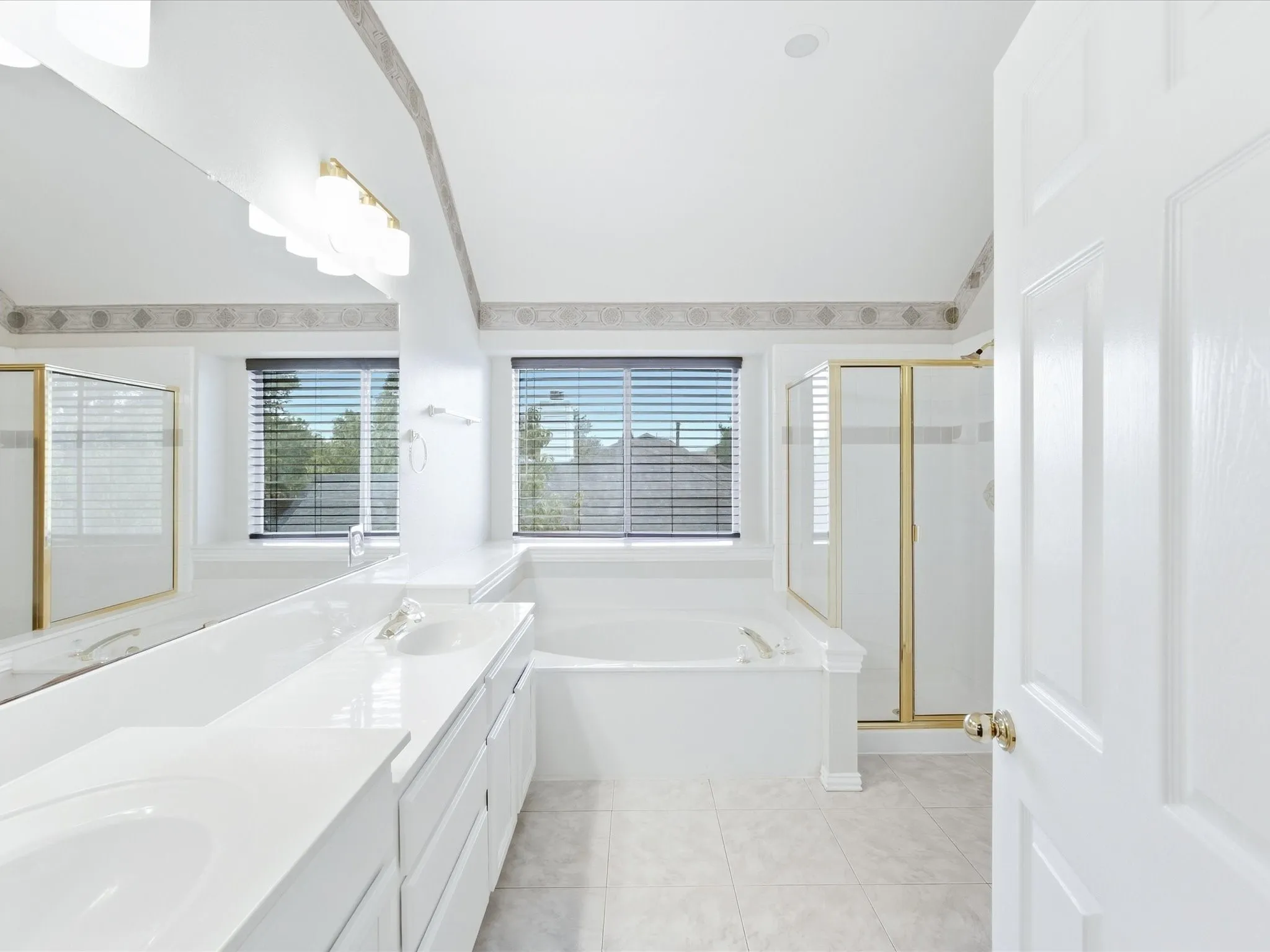Dual vanity with integrated sinks, built-in bathtub, glass-enclosed shower, tiled flooring, and multiple windows with blinds
