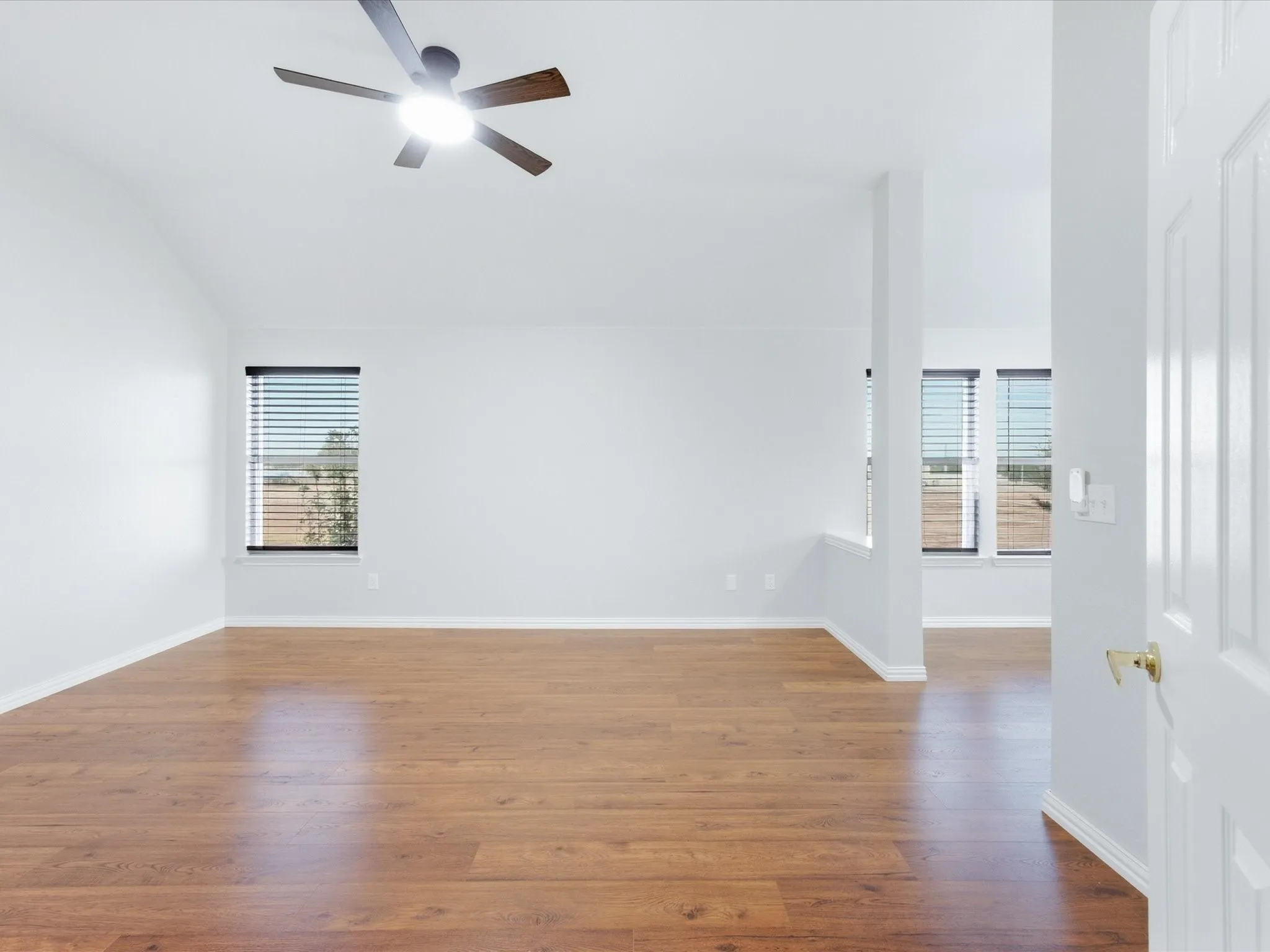 Vaulted ceiling, wood-finish flooring, ceiling fan with integrated lighting, multiple windows with blinds, and white interior paint