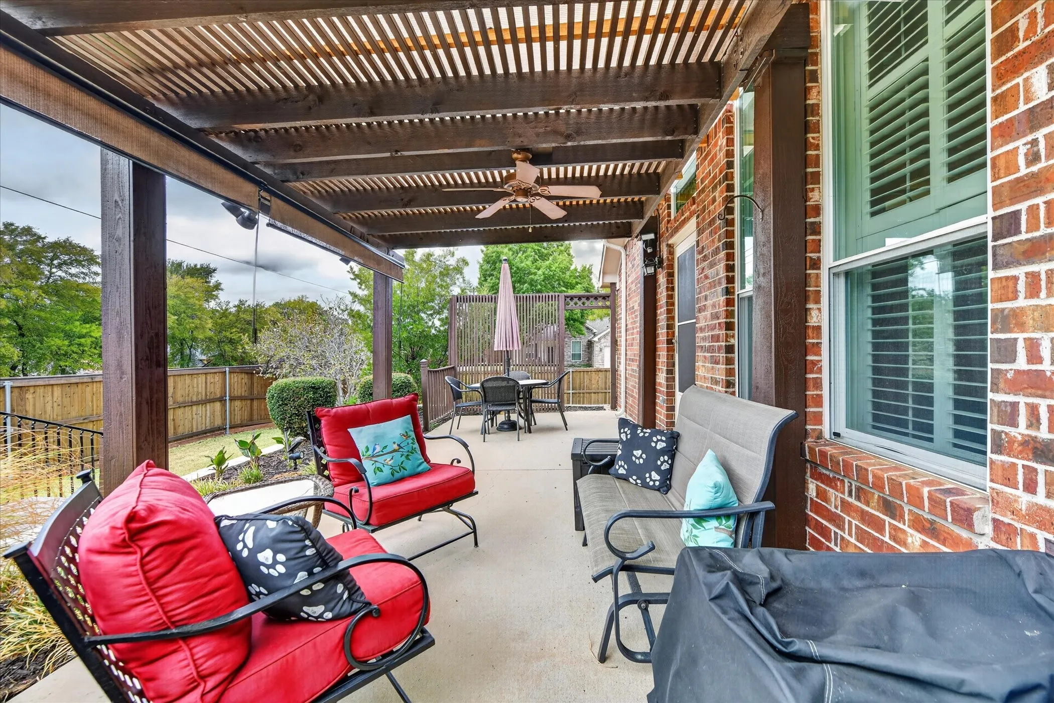 Concrete patio featuring an overhead pergola with a ceiling fan, brick exterior, and white-framed windows with plantation shutters
