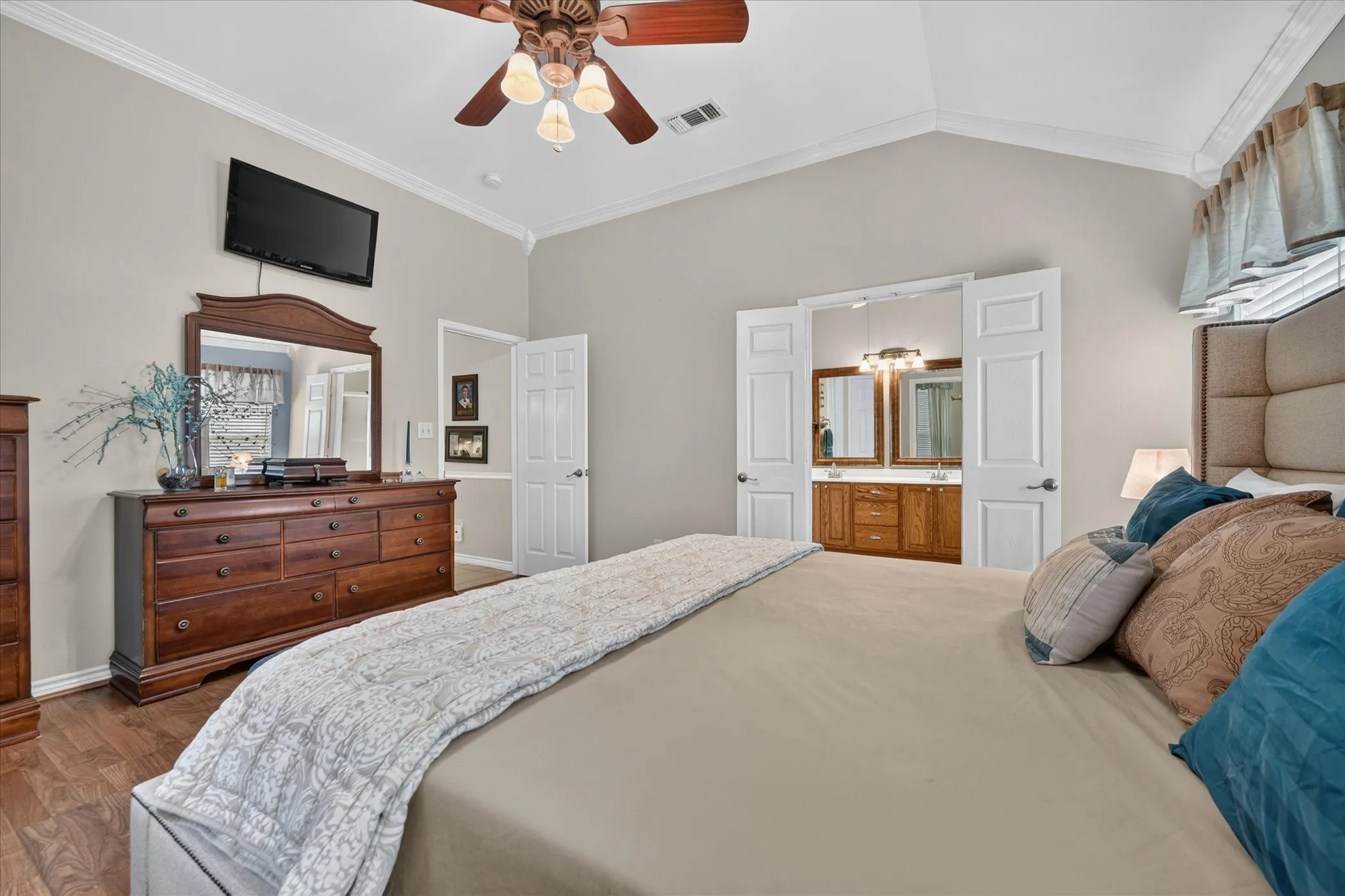 Vaulted ceiling with crown molding, wood-finish flooring, ceiling fan with integrated lighting, dual vanity with wood cabinetry, and framed mirrors