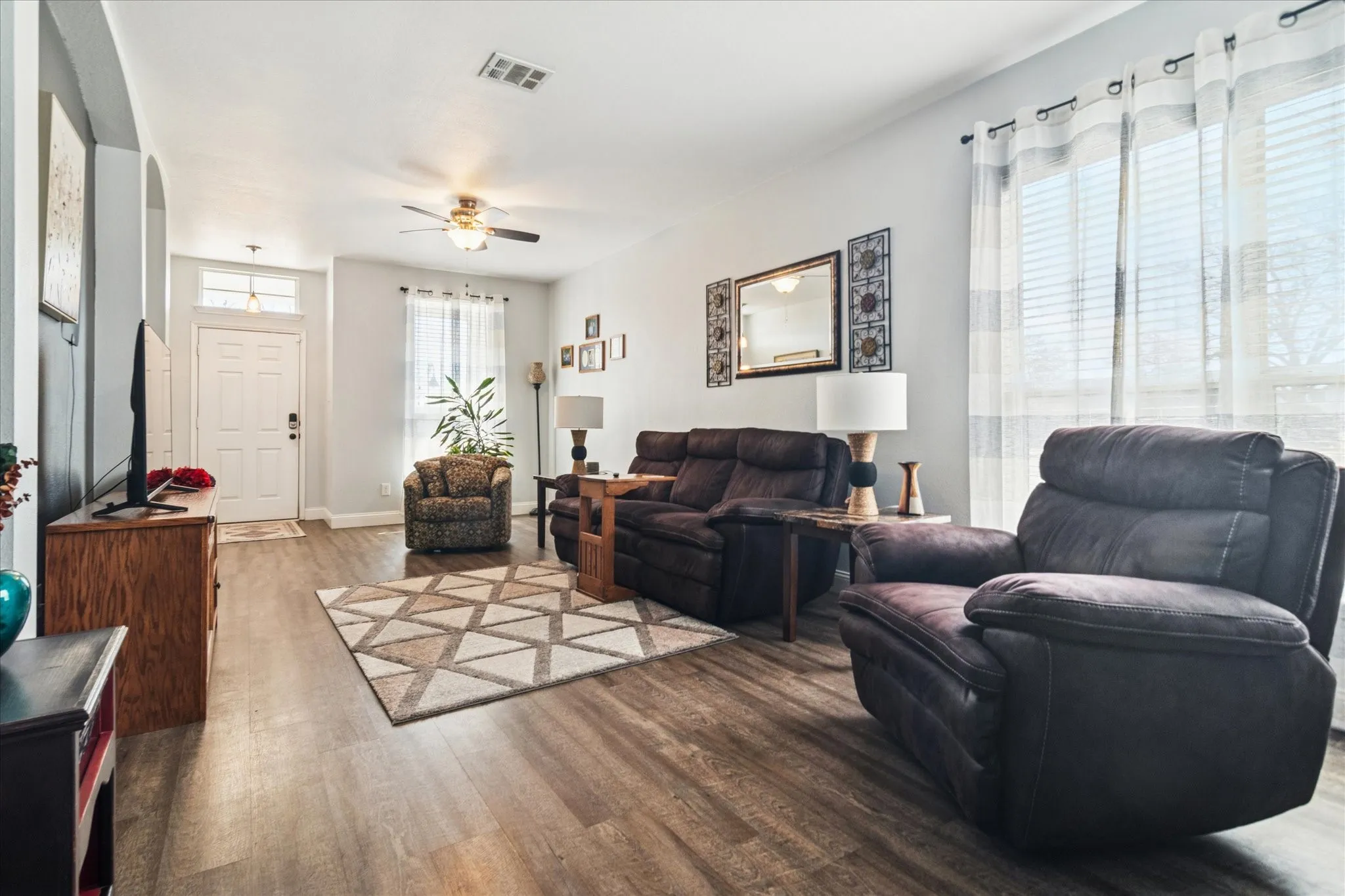Spacious living area featuring wood-finish flooring, a ceiling fan with integrated lighting, and multiple windows with blinds