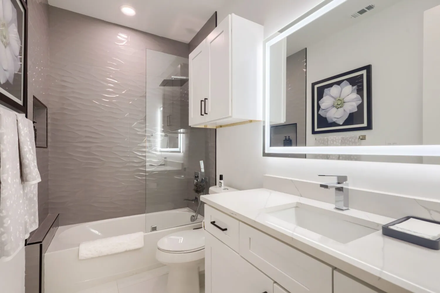 Built-in bathtub with glass shower enclosure, wave-textured tile surround, vanity with integrated sink, chrome faucet, and illuminated mirror