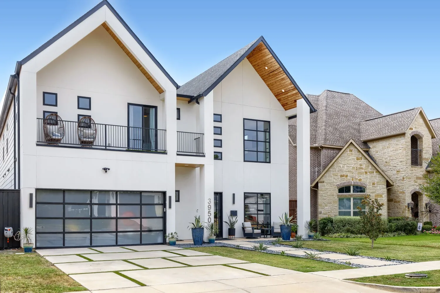 Modern facade with a peaked roofline, wood-finish eaves, a balcony with black railings, a glass panel garage door, and a concrete paver driveway
