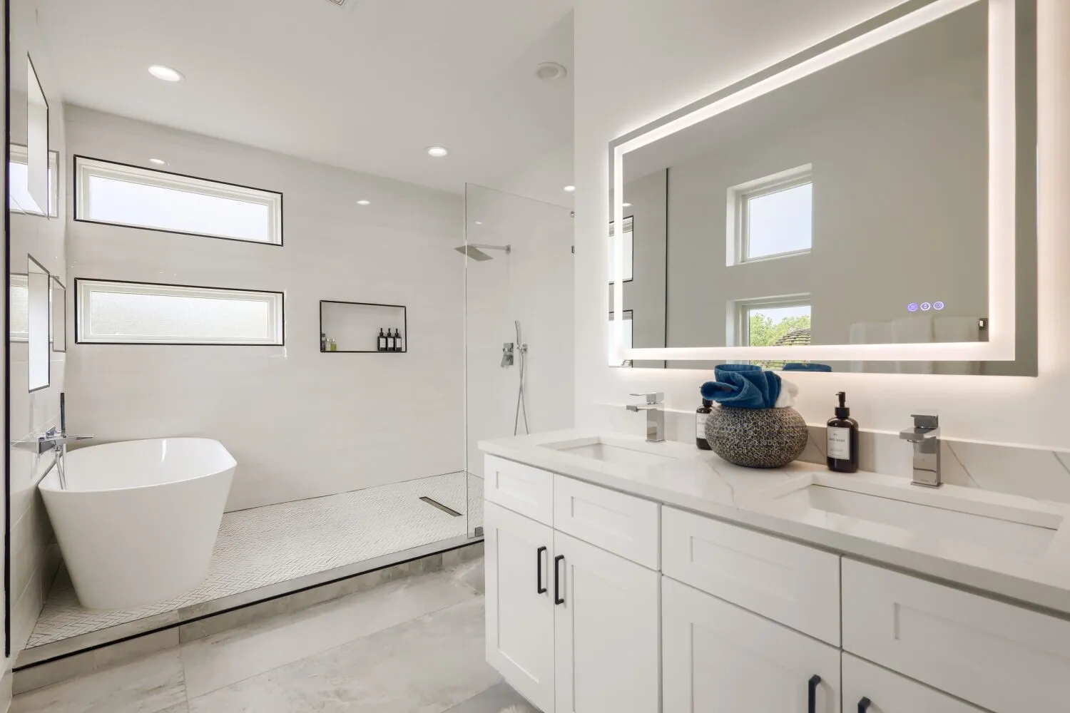 Dual vanity with stone countertops and undermount sinks, illuminated mirror, freestanding soaking tub, glass-enclosed shower, and recessed wall niches