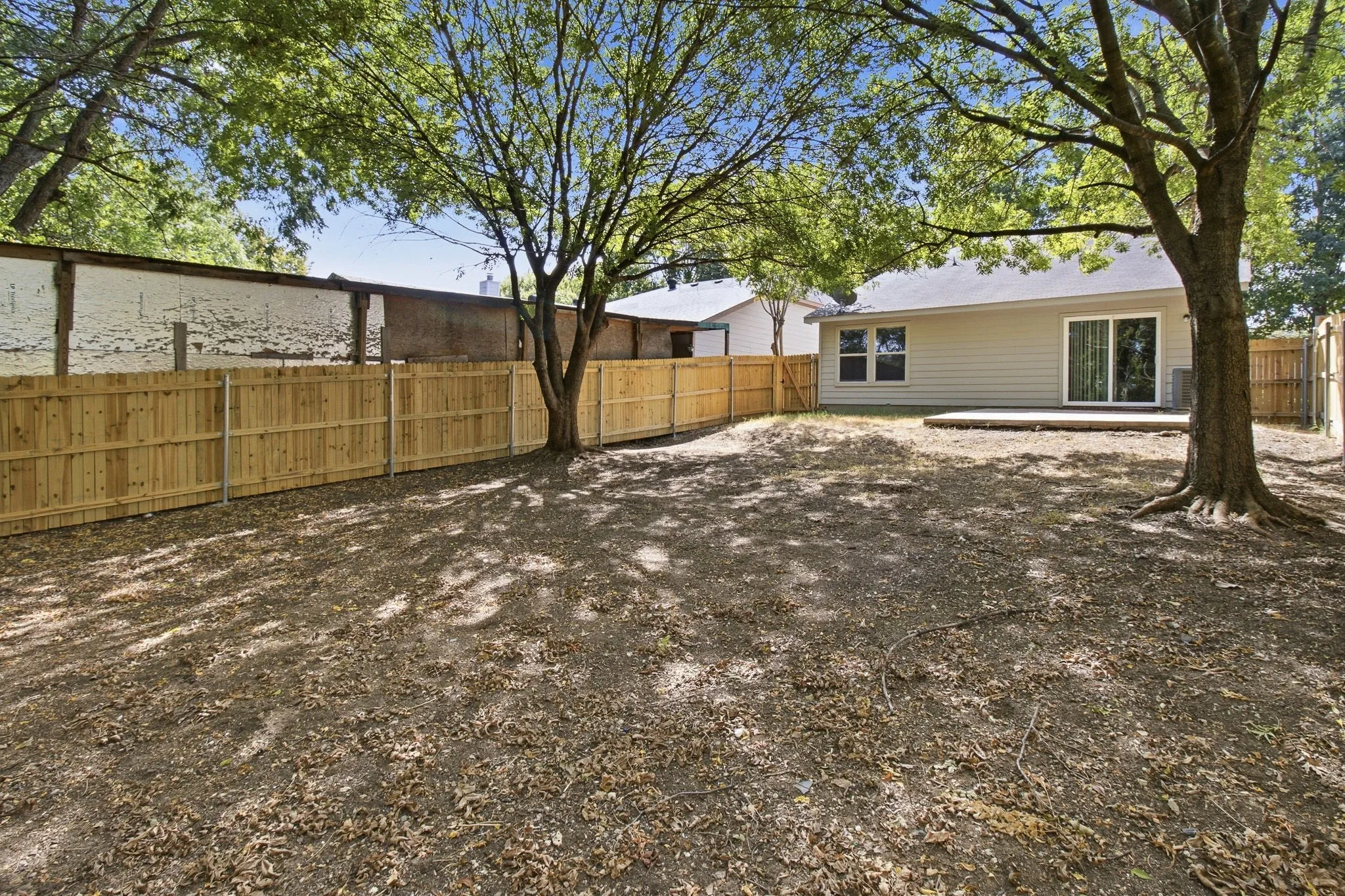 Expansive backyard featuring a wood privacy fence, mature trees, a concrete patio, and a sliding glass door
